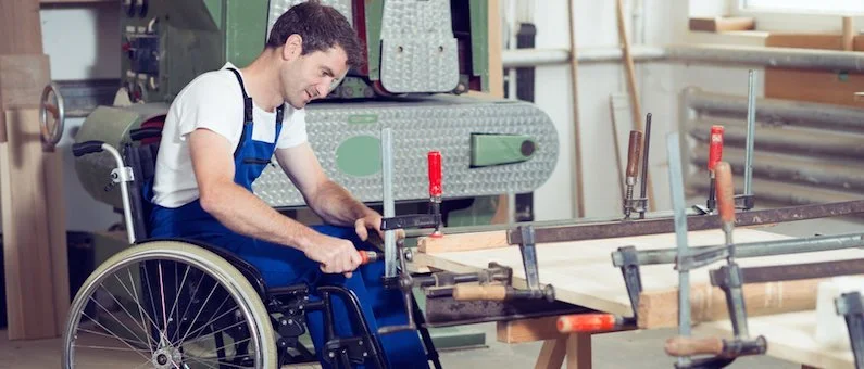 A man in a wheelchair working with woodworking clamps at a workbench in a woodworking shop.
