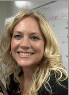 Darcy has curly blonde hair and is smiling in front of a classroom whiteboard.