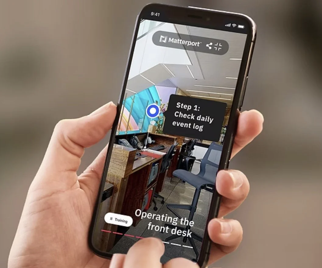 A person holding a smartphone displaying an augmented reality training app with instructions to check a daily event log and operate the front desk in an office environment.