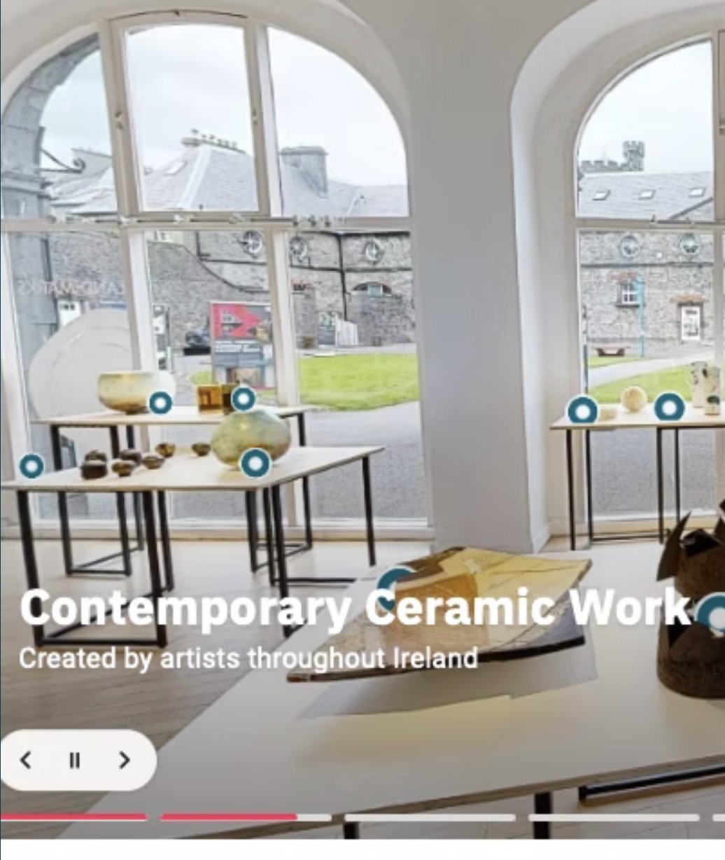 Interior view of an art gallery displaying ceramic artworks on tables near large arched windows with a view of rooftops and green outdoor spaces.