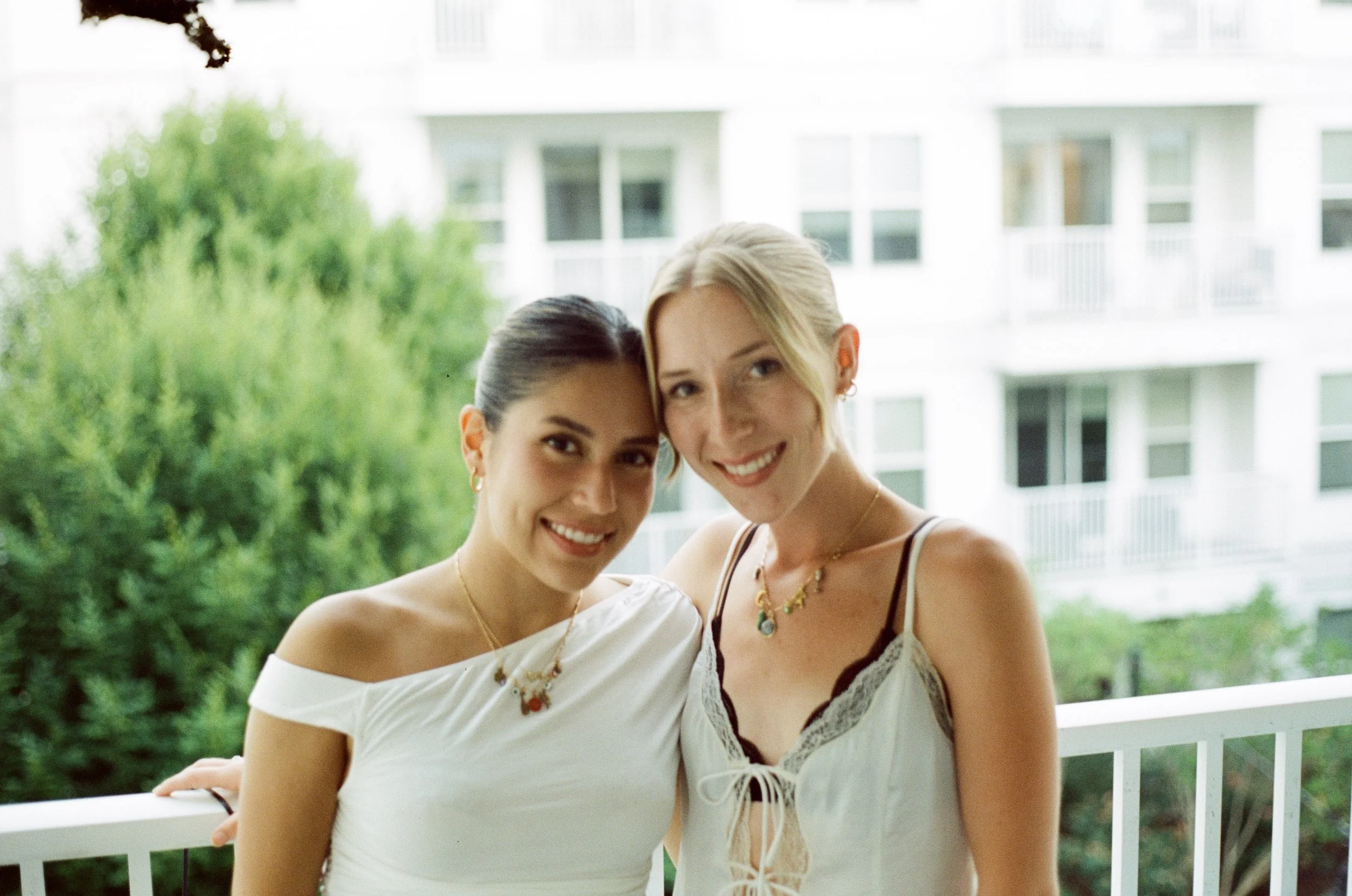 Two smiling women standing close together on a balcony with white railing, a background of green trees and a white apartment building.