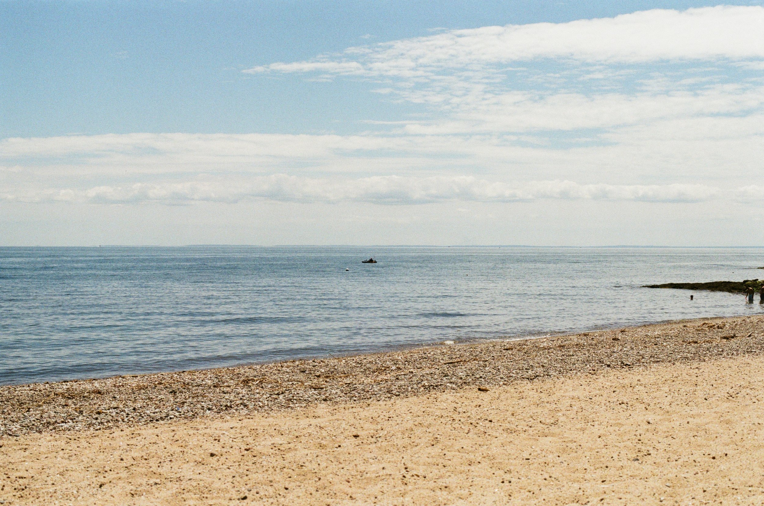 A pebbled beach overlooking calm ocean waters with a small boat in the distance and a partly cloudy sky.