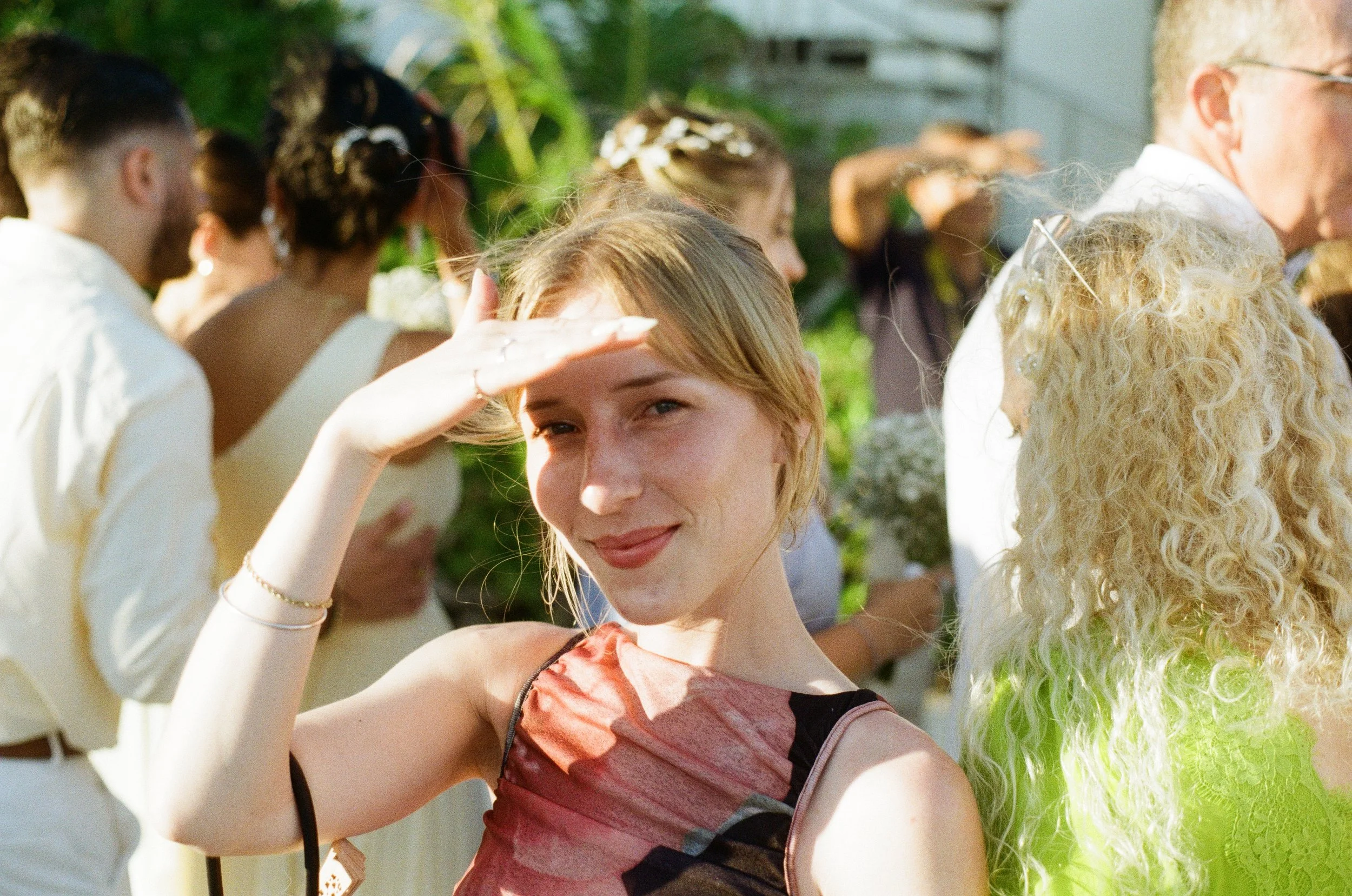 A young woman smiling and shielding her eyes with her hand, standing outdoors among a group of people at a social gathering.