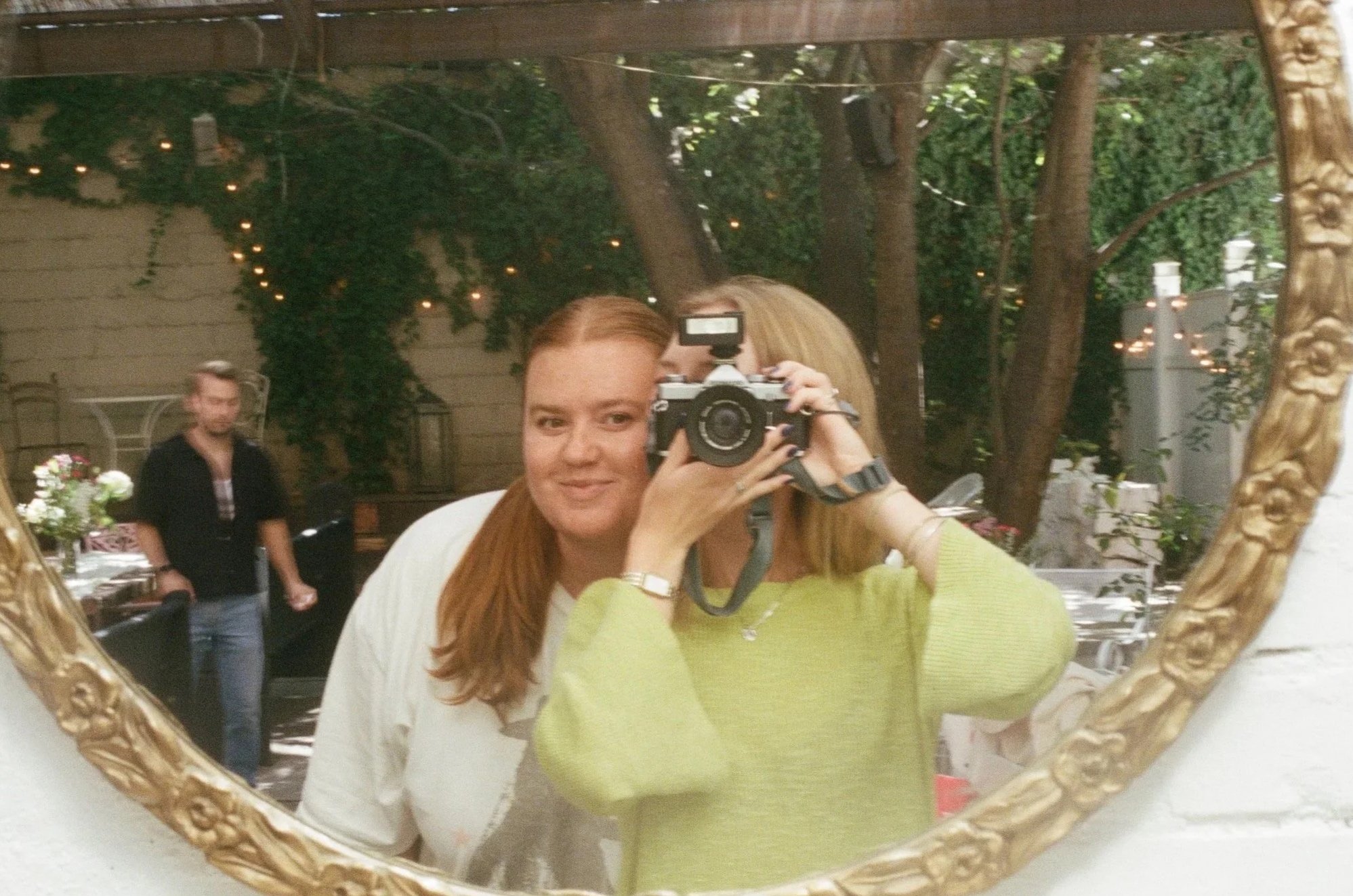 A woman with red hair taking a photo of herself with a camera in a gold ornate mirror. She is wearing a white shirt with a green sweater over it. In the background, a man with blond hair wearing a black shirt is visible, standing near a table with flowers. The setting appears to be an outdoor or semi-outdoor space decorated with string lights and trees.