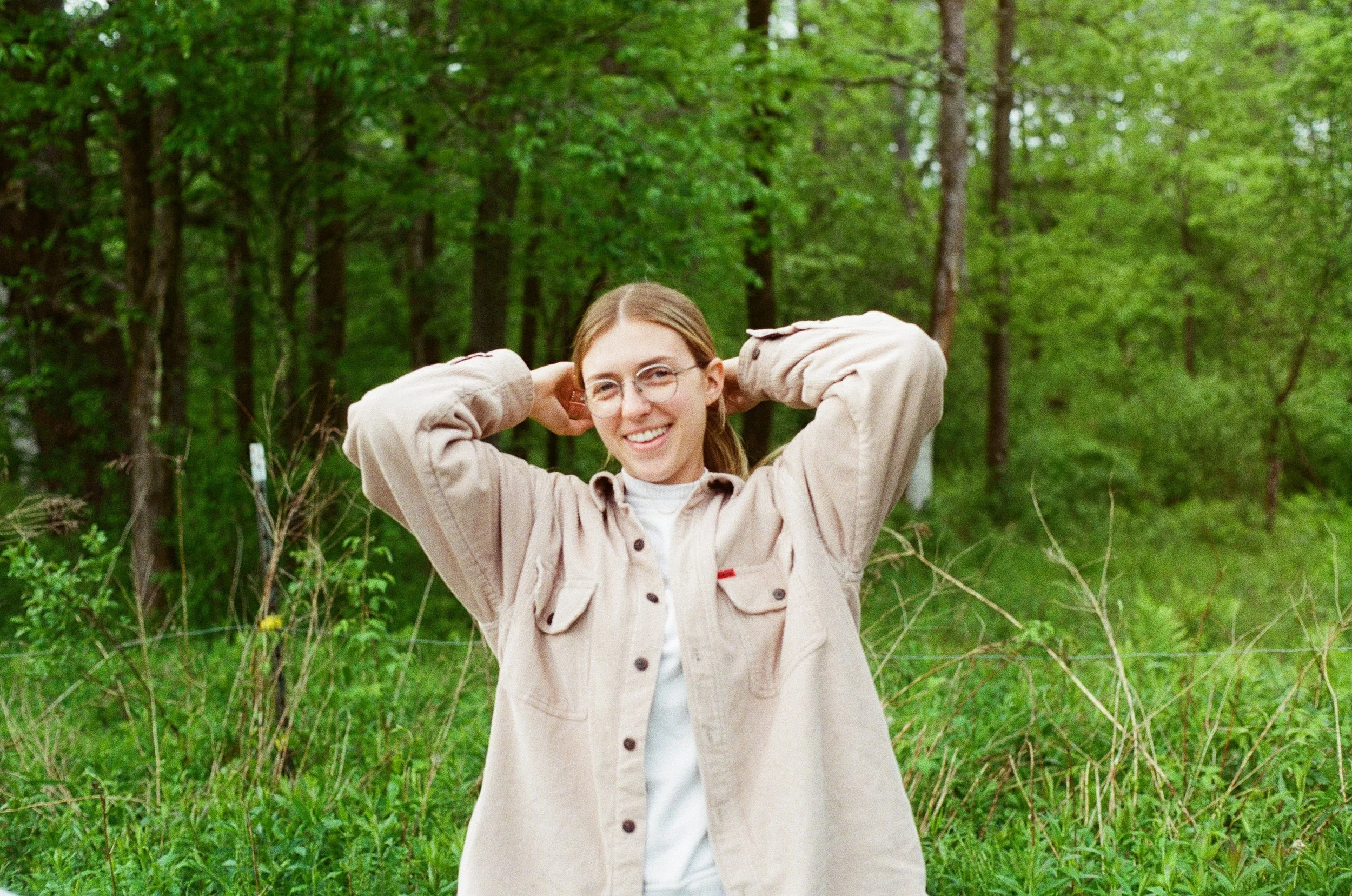 A young woman with glasses and light brown hair, smiling and posing with her hands behind her head, standing in a green forested area.