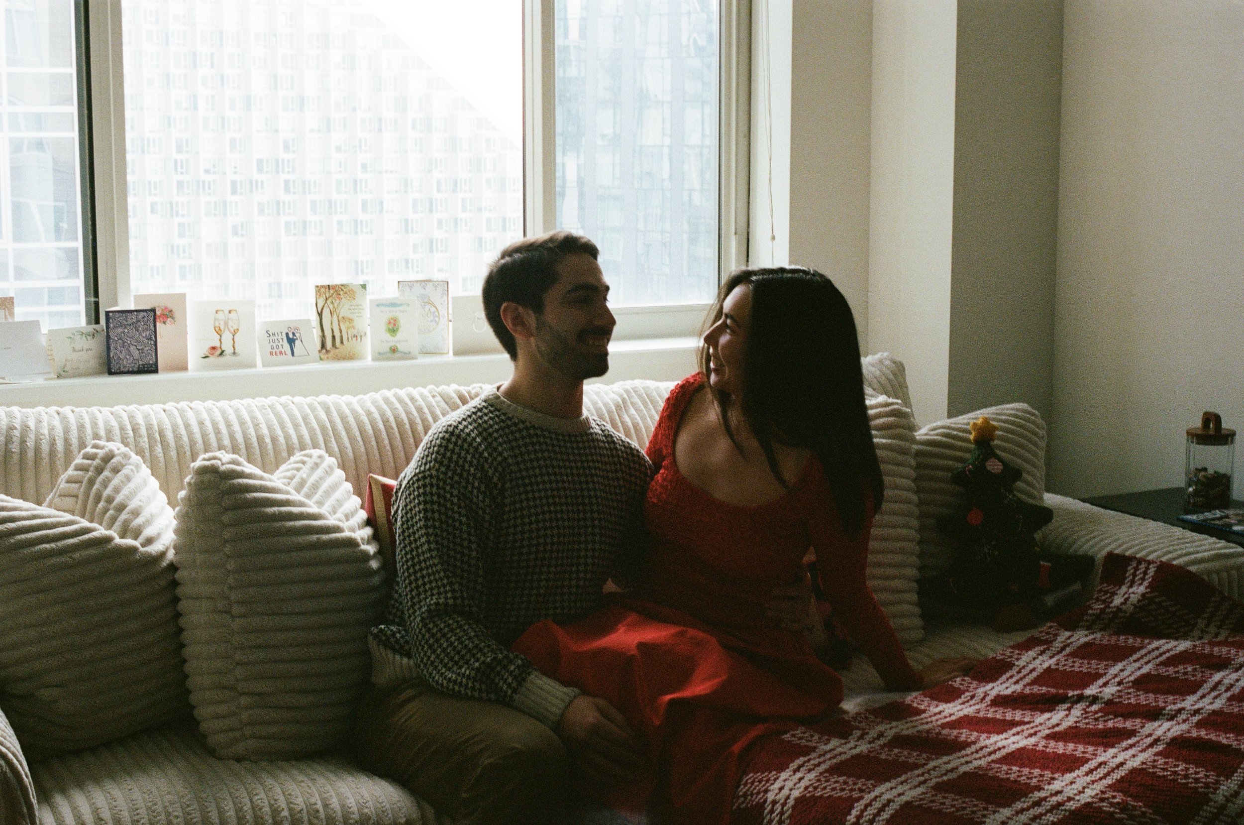A young man and woman sitting close together on a beige striped couch in a living room, smiling and looking at each other, with a large window in the background and holiday cards on the window sill.
