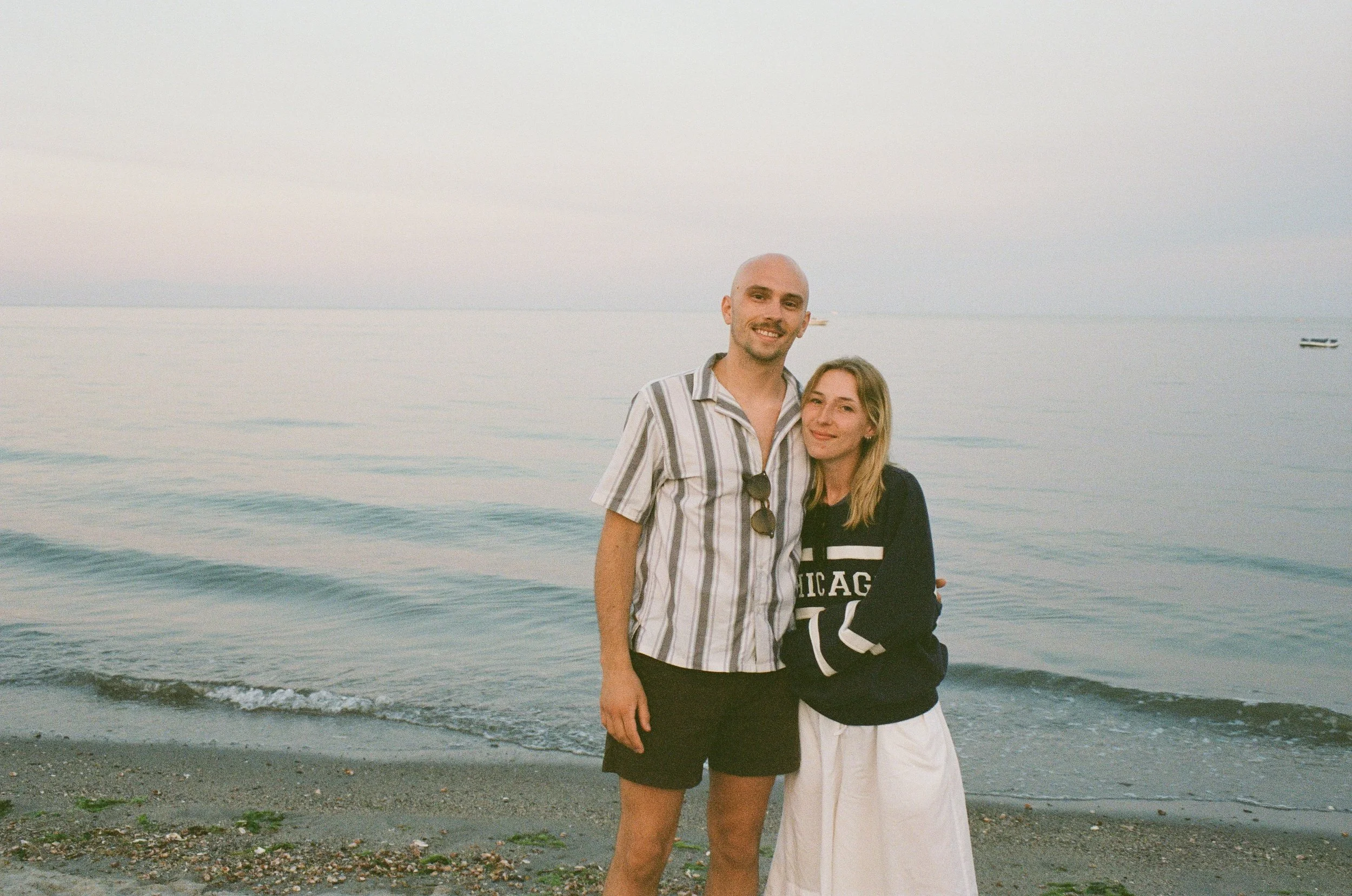 Two people, a man and a woman, standing close together on a beach with water and boats in the background. The man is wearing a striped shirt, and the woman is wearing a black and white sweatshirt with the word 'Chicago' on it.