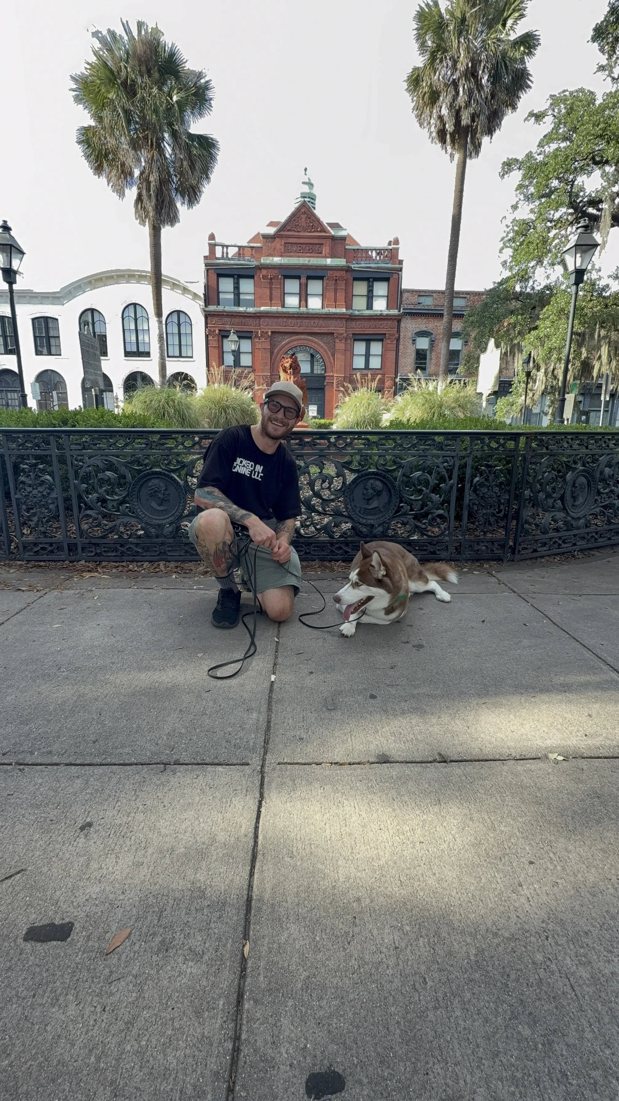 A man with glasses, wearing a black T-shirt and shorts, kneeling on a sidewalk with a brown and white dog lying next to him. Behind them, there is an ornate black fence, lush green plants, and historic brick buildings with palm trees. The man is smiling, and the dog looks relaxed.