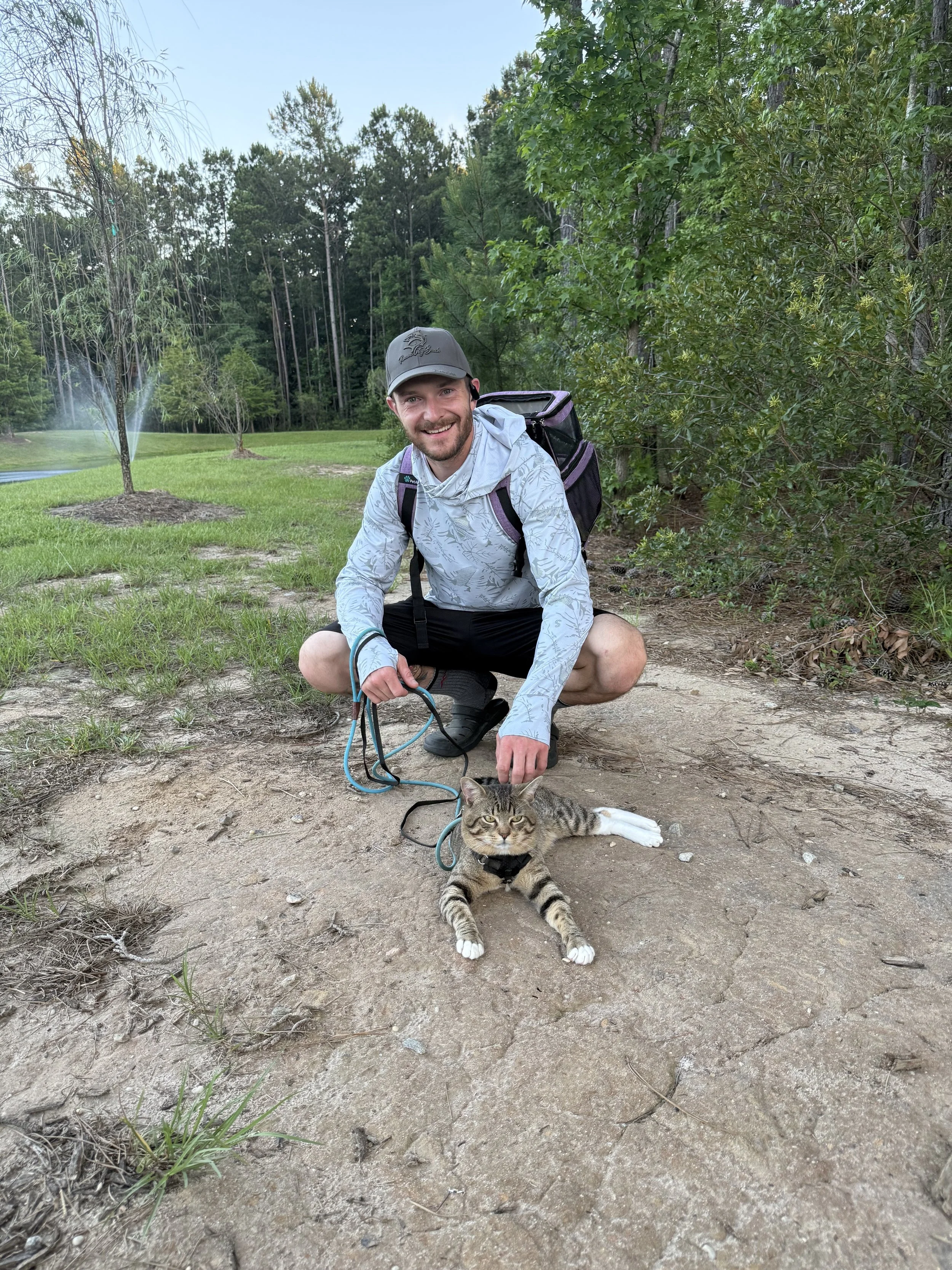 A man squatting outdoors with a backpack, smiling at the camera, while holding a leash attached to a lying tabby cat wearing a harness on a dirt path surrounded by trees and greenery.