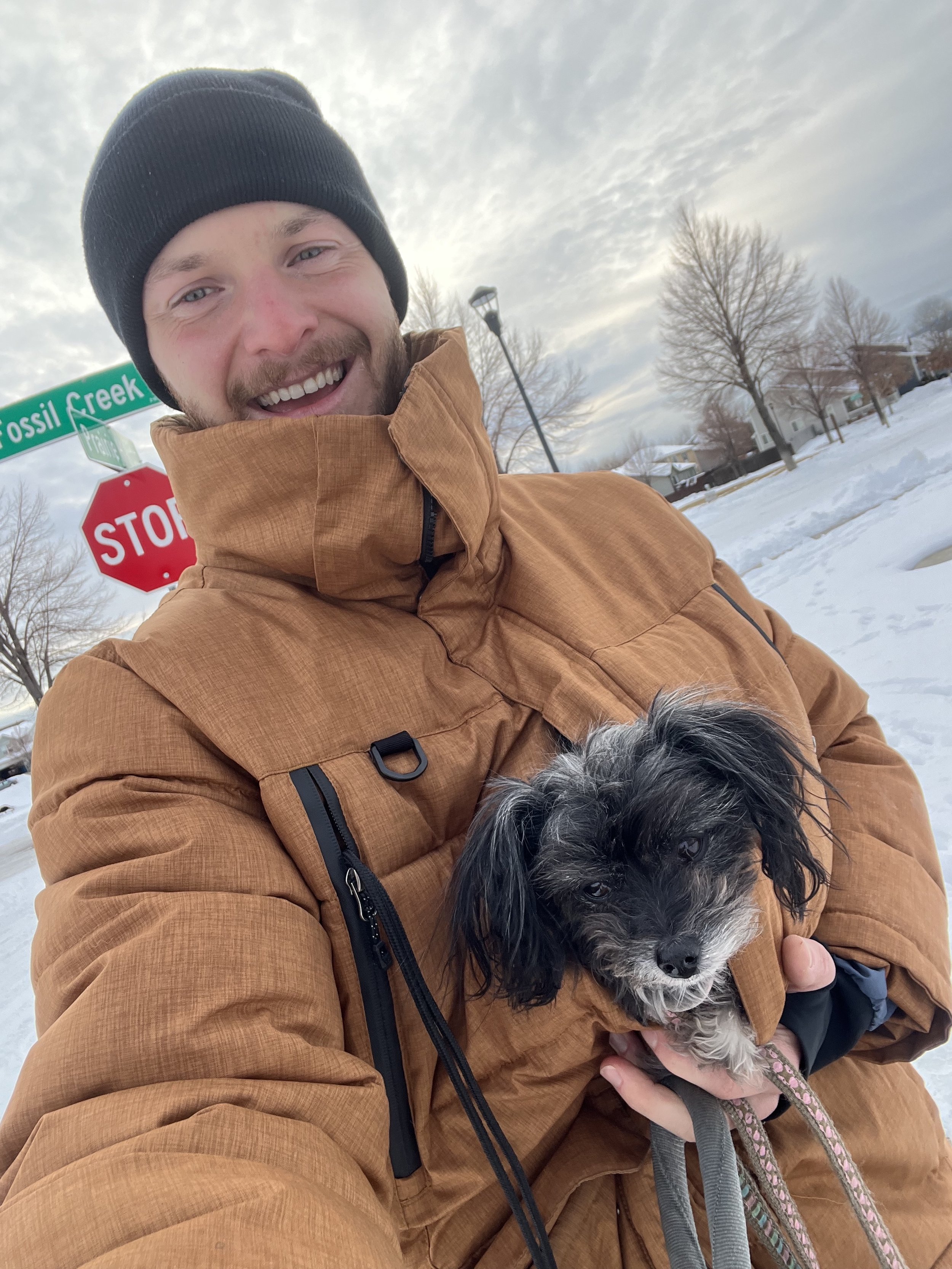 A smiling man in a brown winter jacket and black beanie holding a small black and white dog outside in a snowy neighborhood with leafless trees and houses in the background.