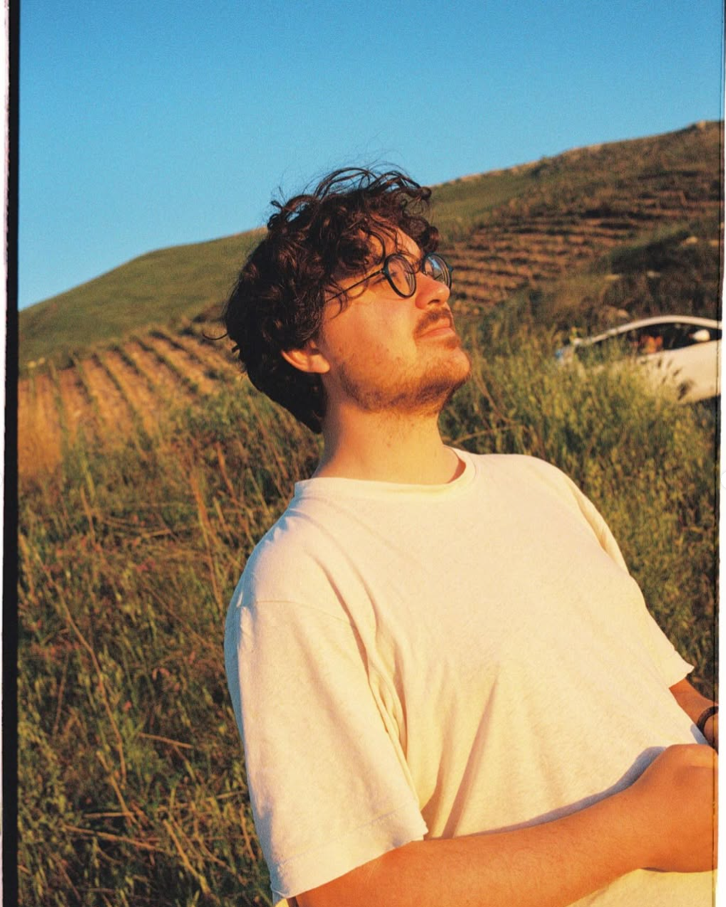 A young man with curly hair and glasses stands outdoors with his eyes closed, enjoying the sunlight. He is wearing a light-colored t-shirt, and the background features a hillside with terraced vegetation and a clear blue sky.