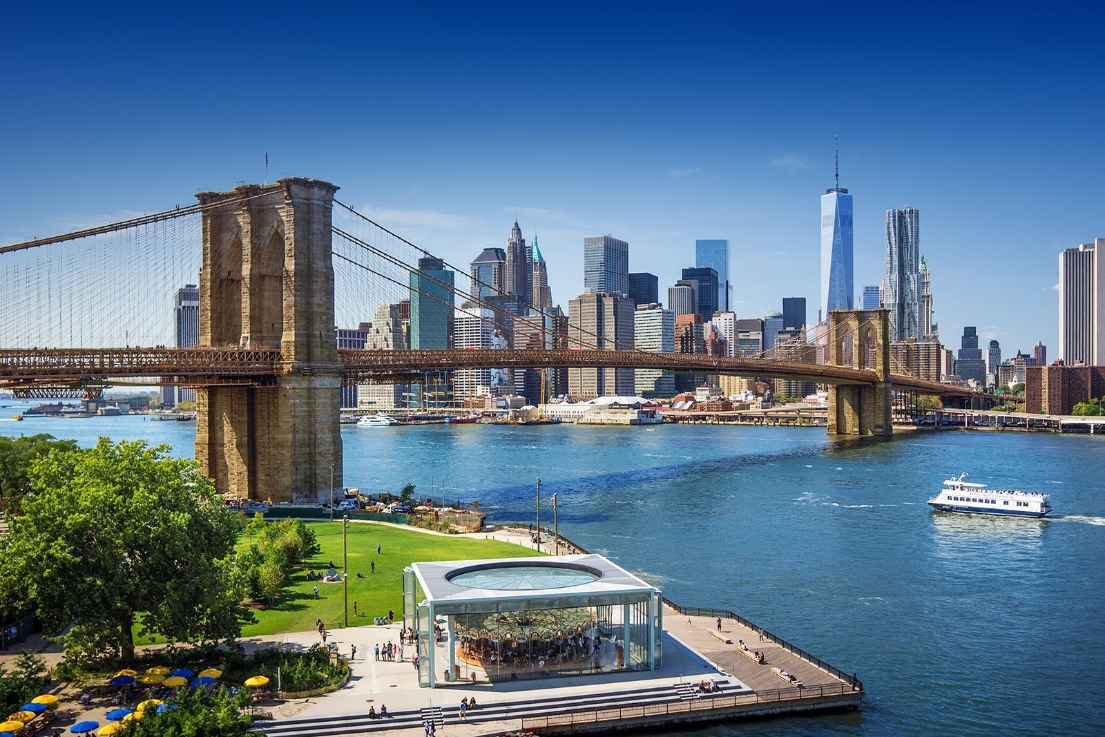 View of the Brooklyn Bridge with Manhattan skyline in the background, a park and a boat on the water in the foreground.