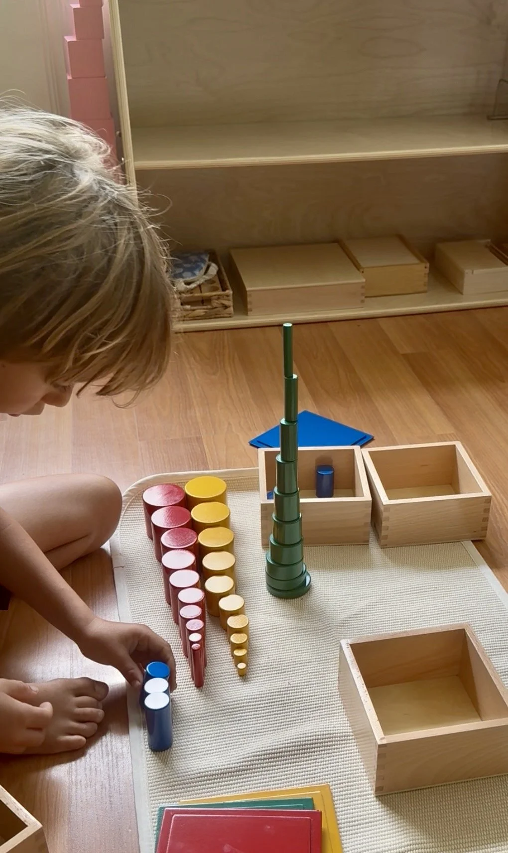 Child playing with colorful stacking and sorting toys on a wooden floor, with empty wooden boxes and shelves in the background.