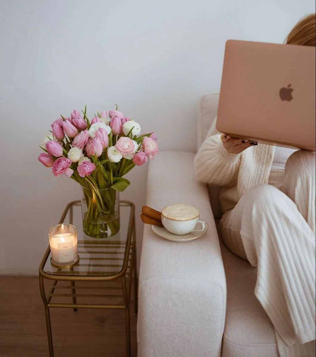 Person using a laptop on a sofa beside a table with pink flowers, a candle, and a cup of coffee.