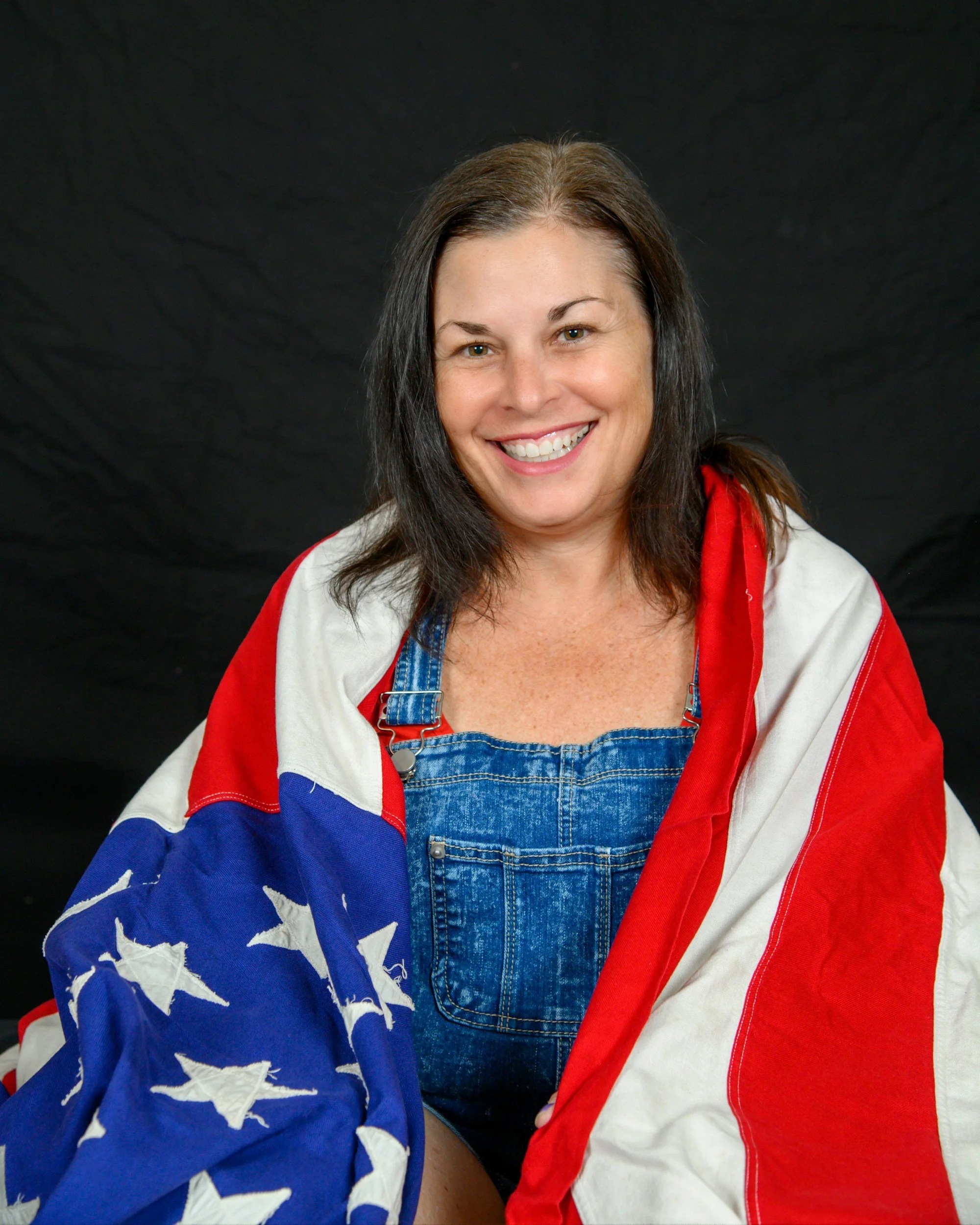 A woman smiling, wearing a jacket with the American flag design, featuring white stars on a blue background, and red and white stripes.