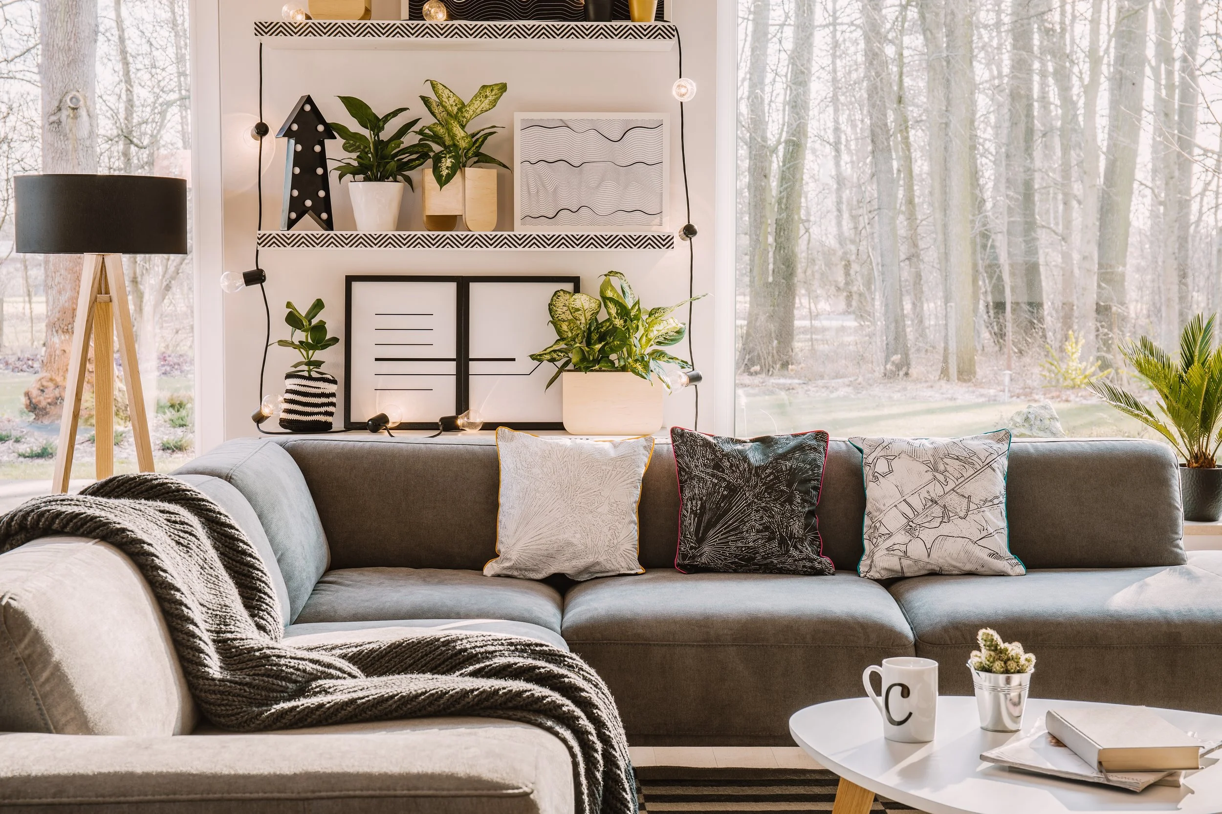 Living room with gray sofa adorned with various patterned pillows, a white round coffee table with books, a cup, and a small pot with plants, and a window with a view of a wooded outdoors area. Decorative elements on the wall include framed art, potted plants, a black star-shaped decor with lights, and string lights.