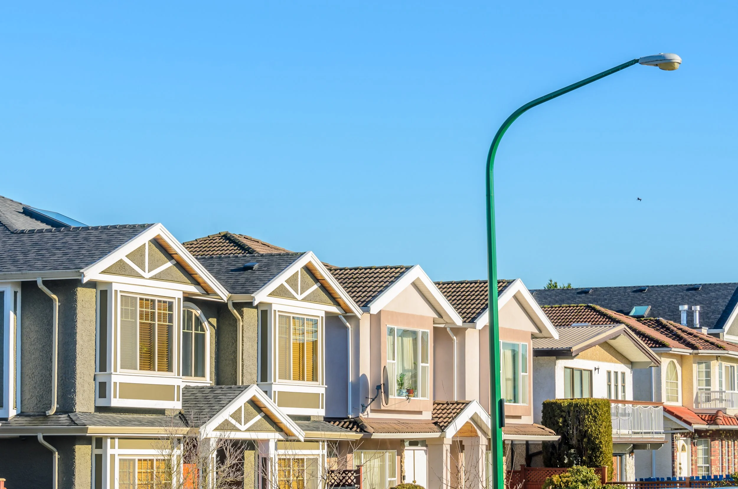 Row of modern suburban houses with various roof styles, a green street lamp, a clear blue sky, and a small bird flying in the background.