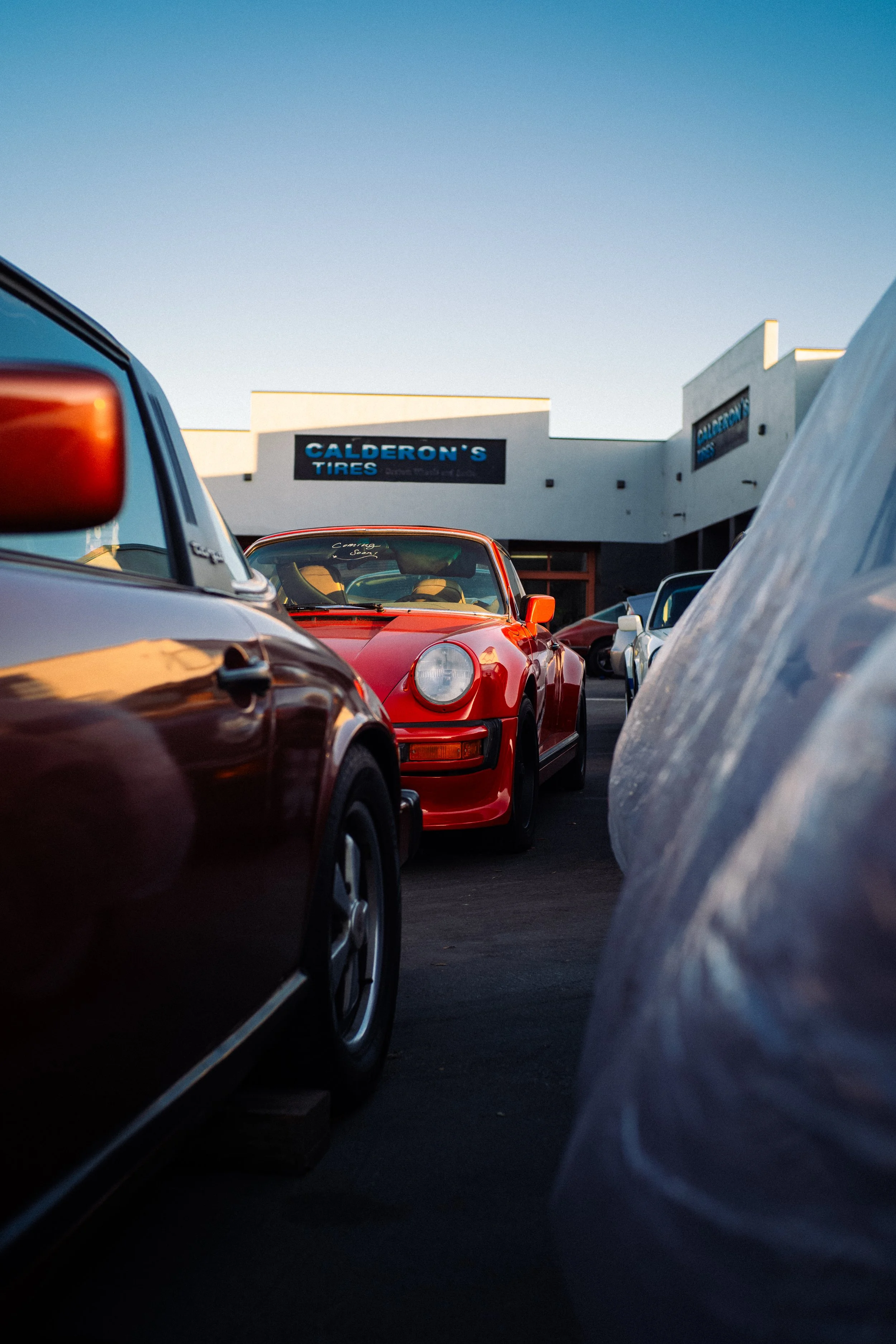 Fotografie von Simon De Luigi, Aufnahmen eines Parkplatzes mit mehreren Autos, darunter ein roter Porsche 911 Oldtimer, im Hintergrund ein Gebäude mit einem Schild "Calderon’s Tires" bei Sonnenuntergang.