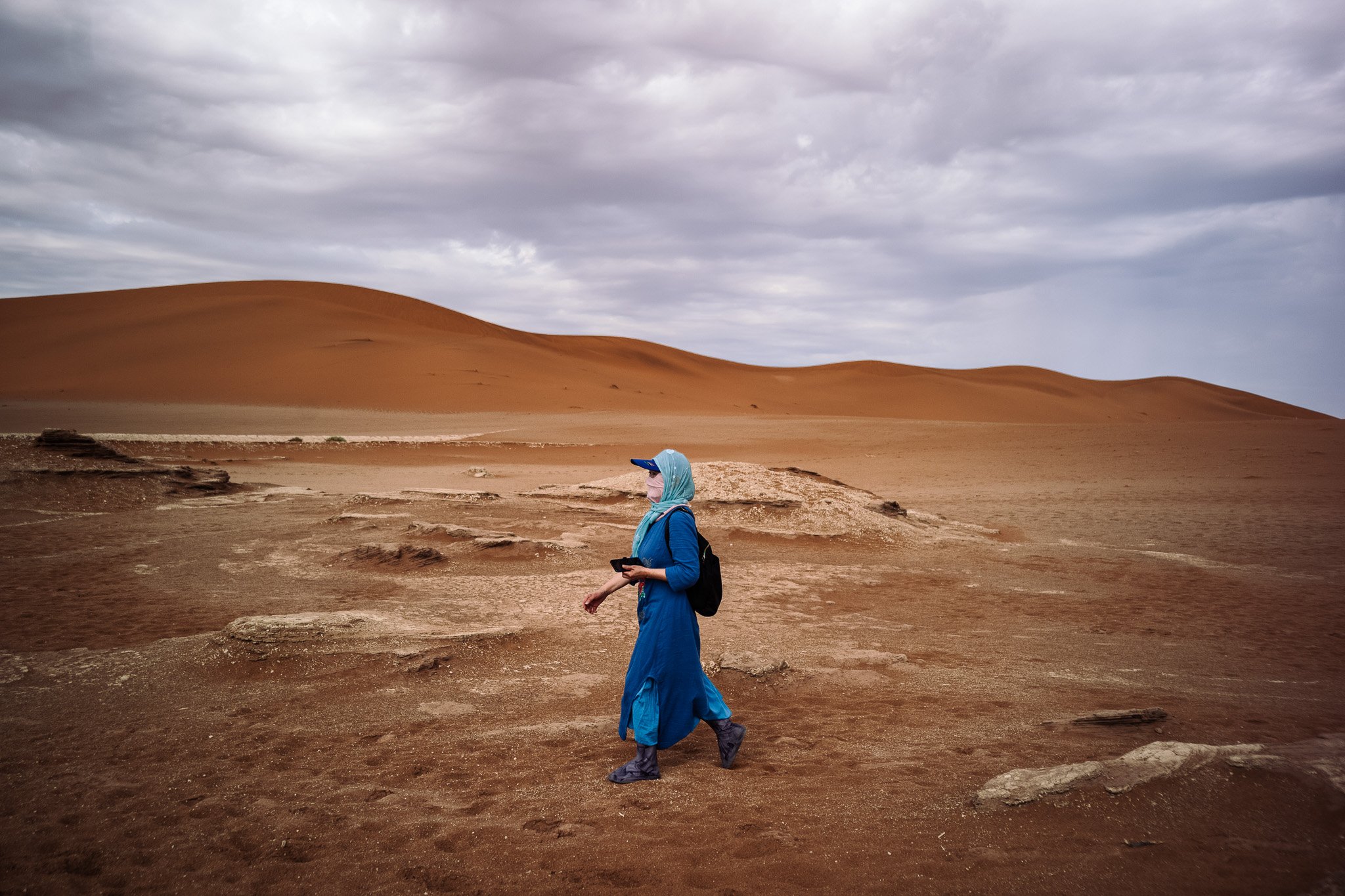 Fotografie von Simon De Luigi, Eine Person in blauer Kleidung, mit Kopftuch und Rucksack, läuft in einer Wüstenlandschaft Namib Wüste unter einem bewölkten Himmel.