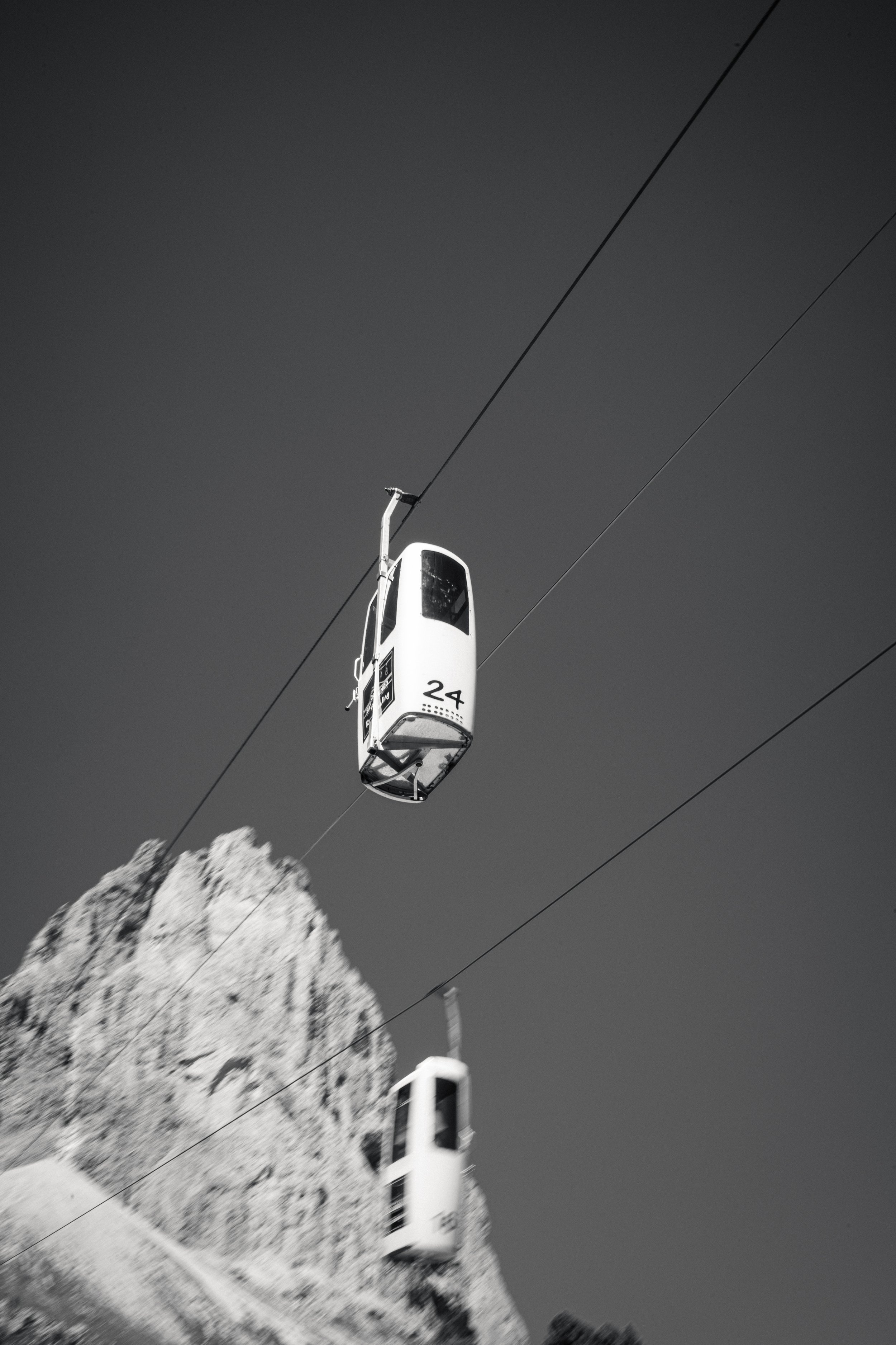 Fotografie von Simon De Luigi, Seilbahn mit Kabine in den Dolomiten, die in den Himmel über einem schneebedeckten Berg fährt, schwarz-weiß