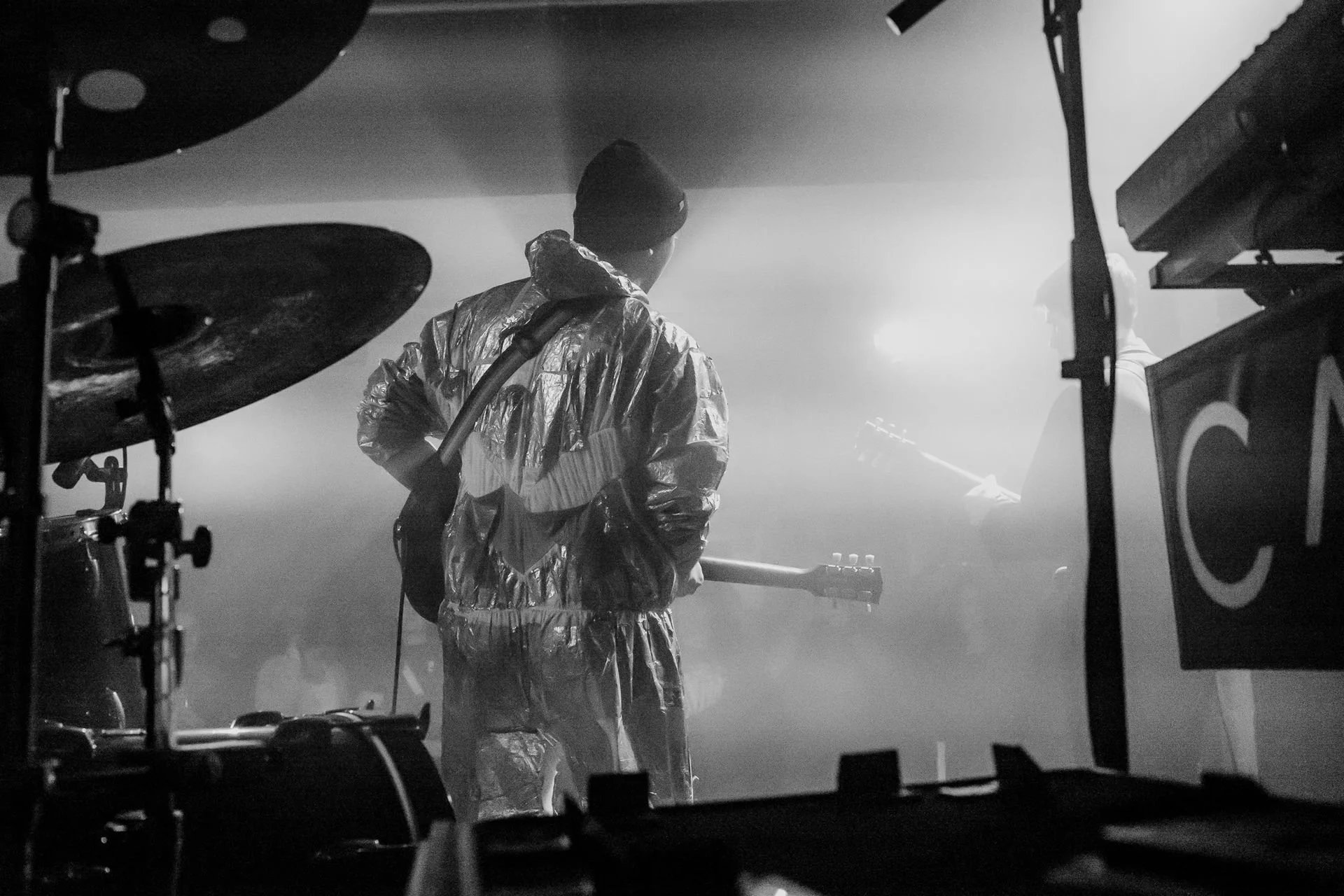 Fotografie von Simon De Luigi, Ein Musiker mit dunkler Mütze und reflektierender Jacke, der eine E-Gitarre spielt, zu sehen durch eine Kameraaufnahme auf einer Bühne mit Rauch und Licht im Hintergrund.