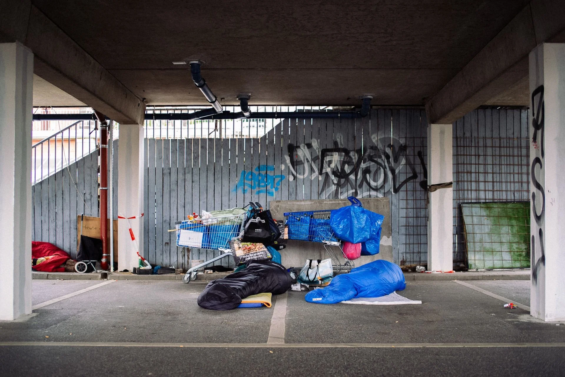 Fotografie von Simon De Luigi, Überdachte Parklücke mit herumliegenden Gegenständen, einschließlich Einkaufswagen, Umlaufdecken und Müllsäcken, sowie Graffiti an der Wand im Hintergrund.