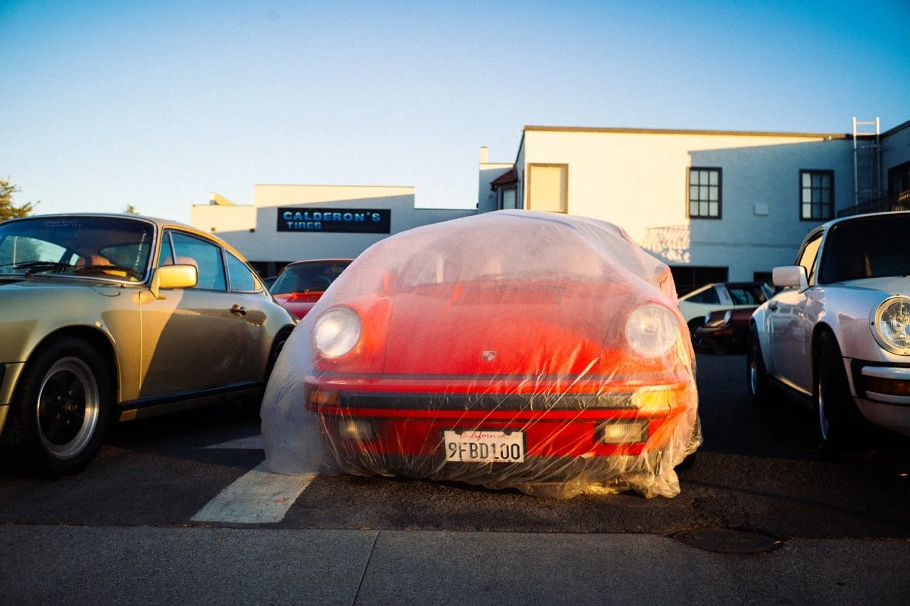 Fotografie von Simon De Luigi, roter Porsche 911 Oldtimer, eingewickelt in eine Plastikhülle, parkiert zwischen anderen Fahrzeugen in einem Parkplatz bei Sonnenuntergang