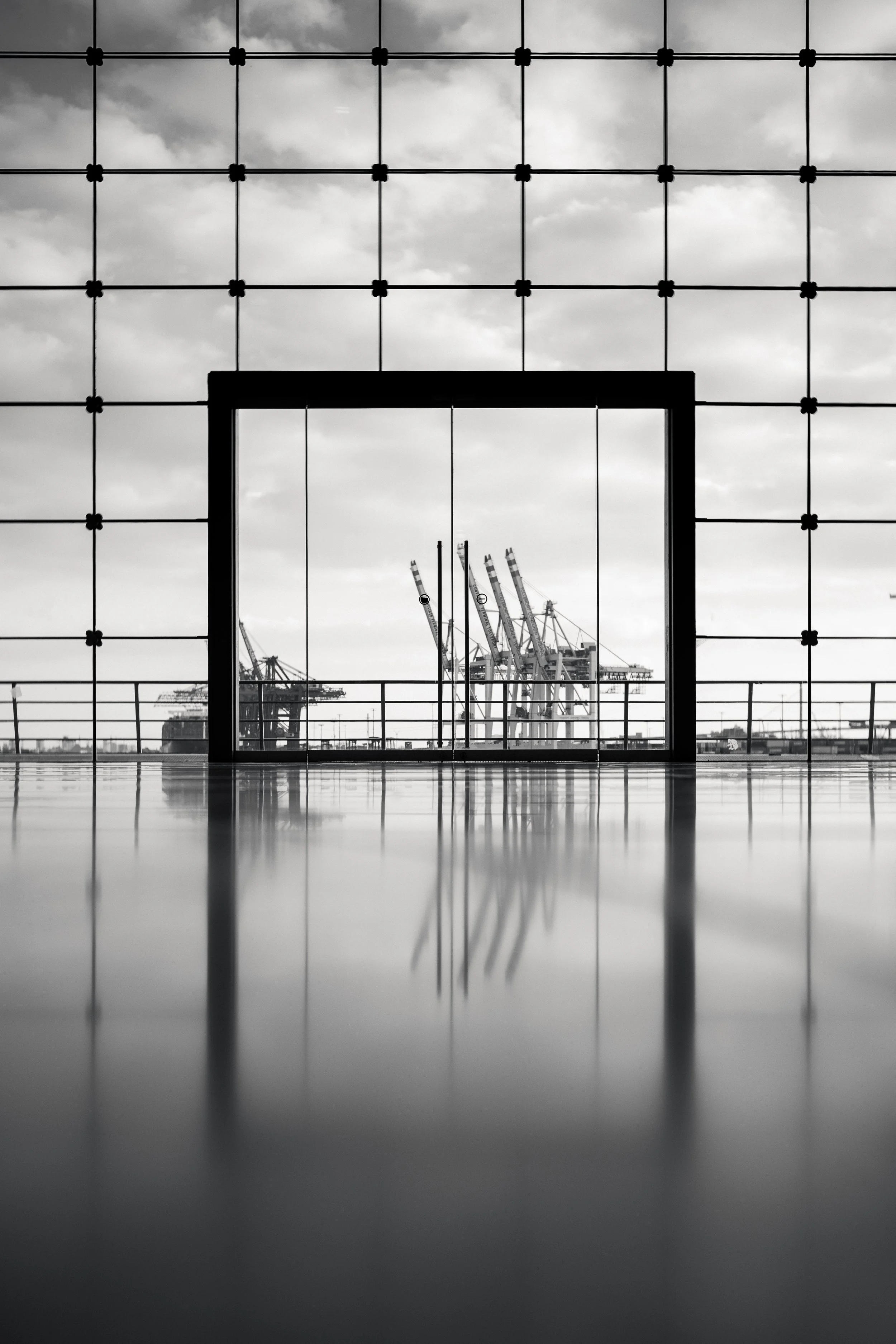 Fotografie von Simon De Luigi, Schwarz-Weiß-Foto eines Gebäudes am Hamburger Fischmarkt, mit einer großen Fensterfront im Vordergrund, durch die man auf eine Industriestadt mit Kranen und Fabrikschornsteinen im Hintergrund sieht.
