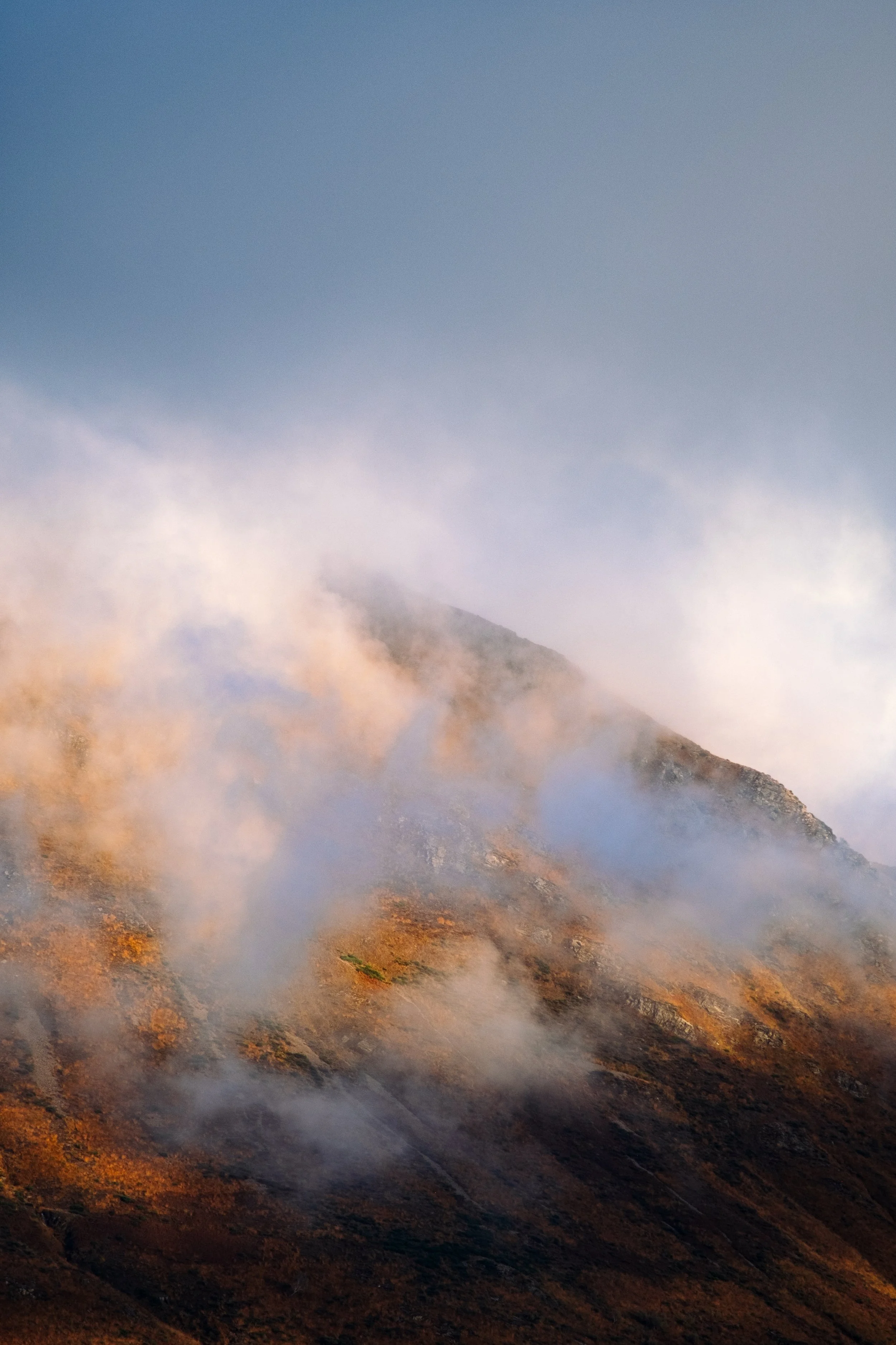 Fotografie von Simon De Luigi, Berggipfel im Tessin, eingehüllt in einer Wolke, mit einer warmen, felsigen Landschaft im Vordergrund.