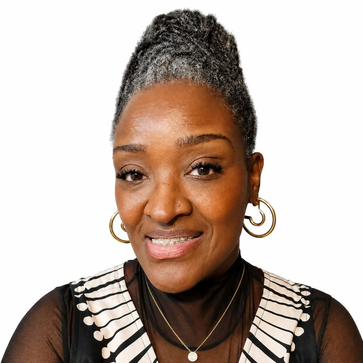 Close-up of a smiling Black woman with curly gray hair styled in an updo, wearing gold hoop earrings, a striped top with sheer black panels, and layered necklaces, against a white background.