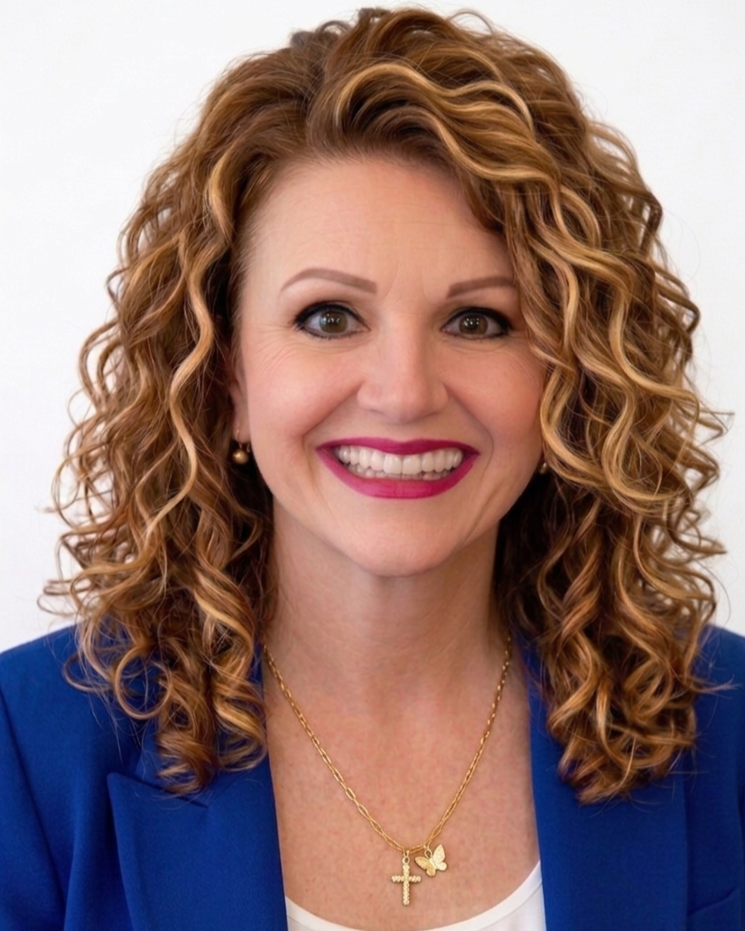 A woman with curly, reddish hair wearing a blue blazer, gold jewelry, and bright pink lipstick, smiling at the camera.