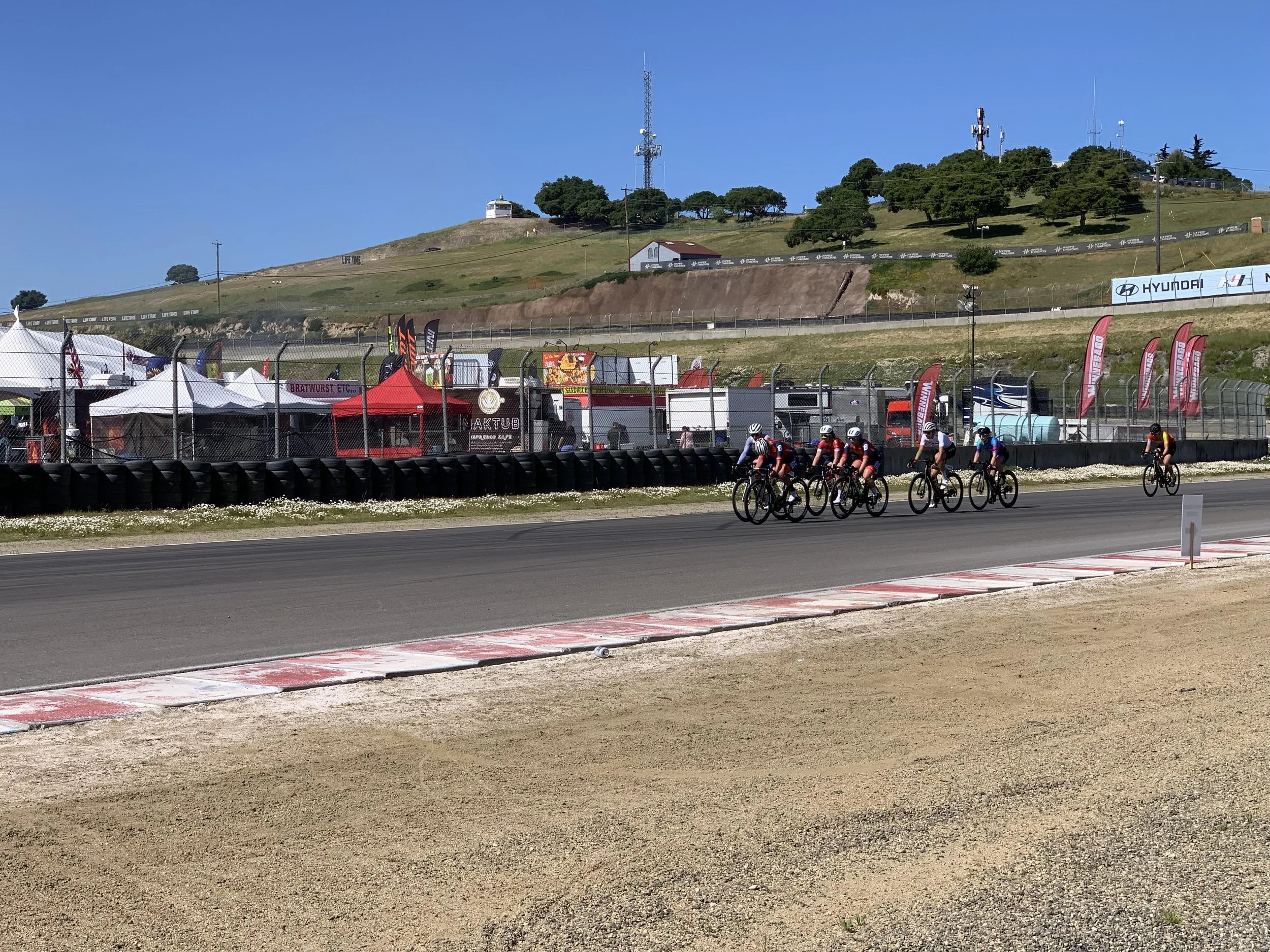 Cyclists riding on a race track with tents and flags in the foreground, a hill with trees and communication towers in the background, under a clear blue sky.