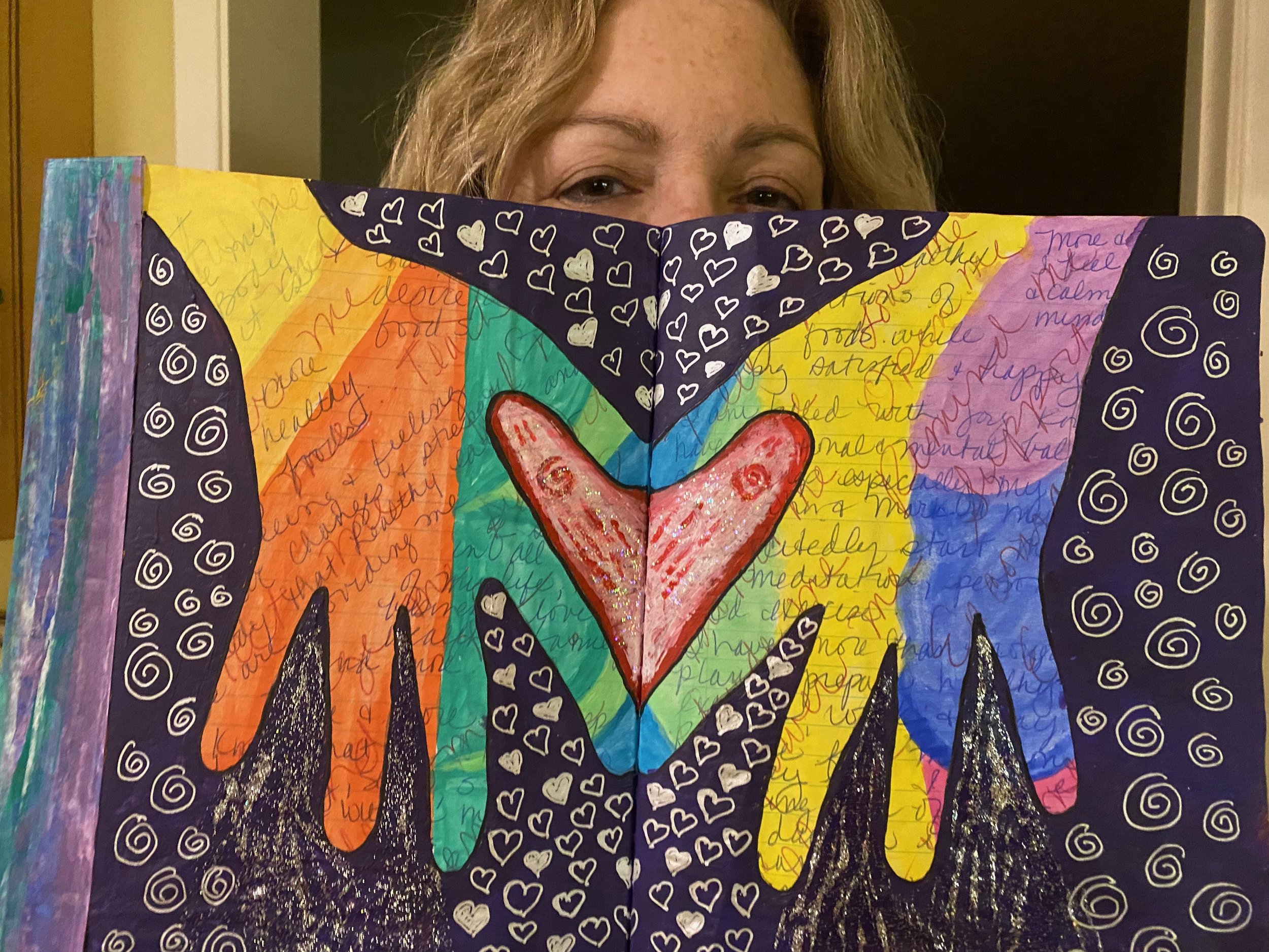 Colorful drawing on paper with a rainbow heart at the center, surrounded by handwritten words and decorated with various patterns including hearts, swirls, and black-and-white sections. A person peeks from behind the artwork.