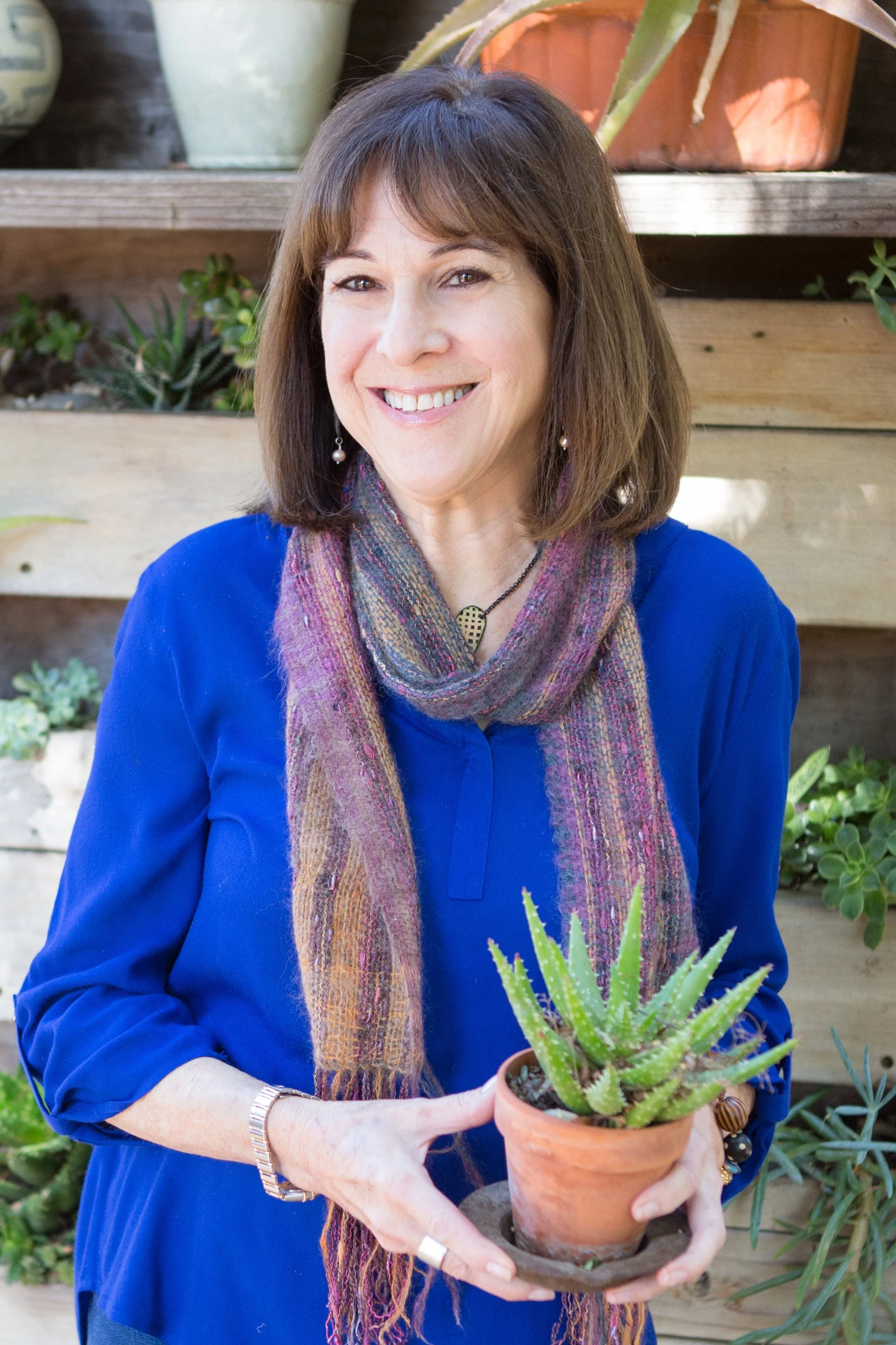 Nancy Chaconas in a blue shirt holding a potted succulent plant, standing in front of a wooden wall with plants and decorative pots.