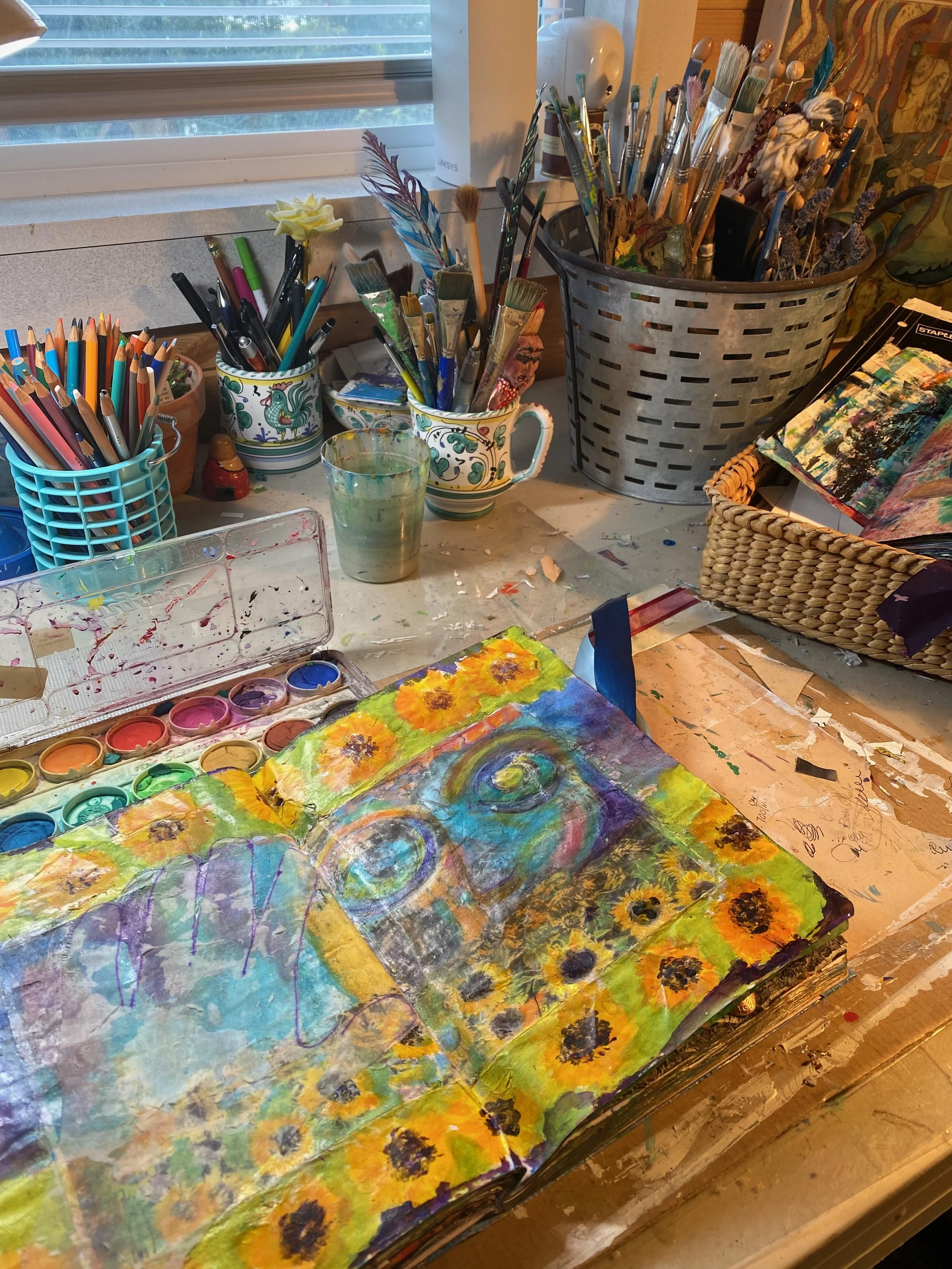 A cluttered artist's workspace with paint brushes, colored pencils, a cup of water, and an abstract painting of sunflowers.