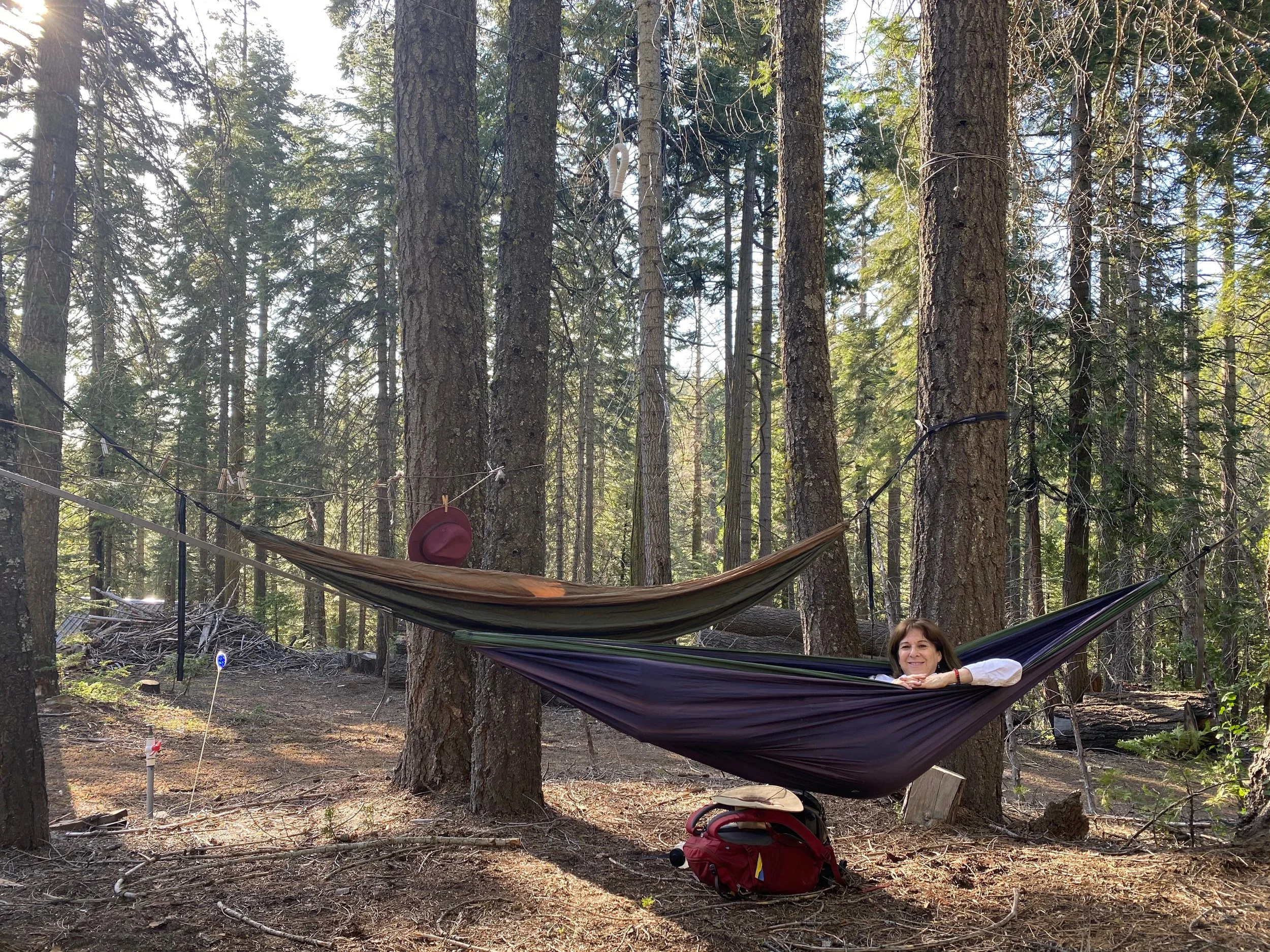 Woman lying in a hammock between trees in a forest, with a red backpack and hat on the ground nearby and sunlight filtering through the trees.
