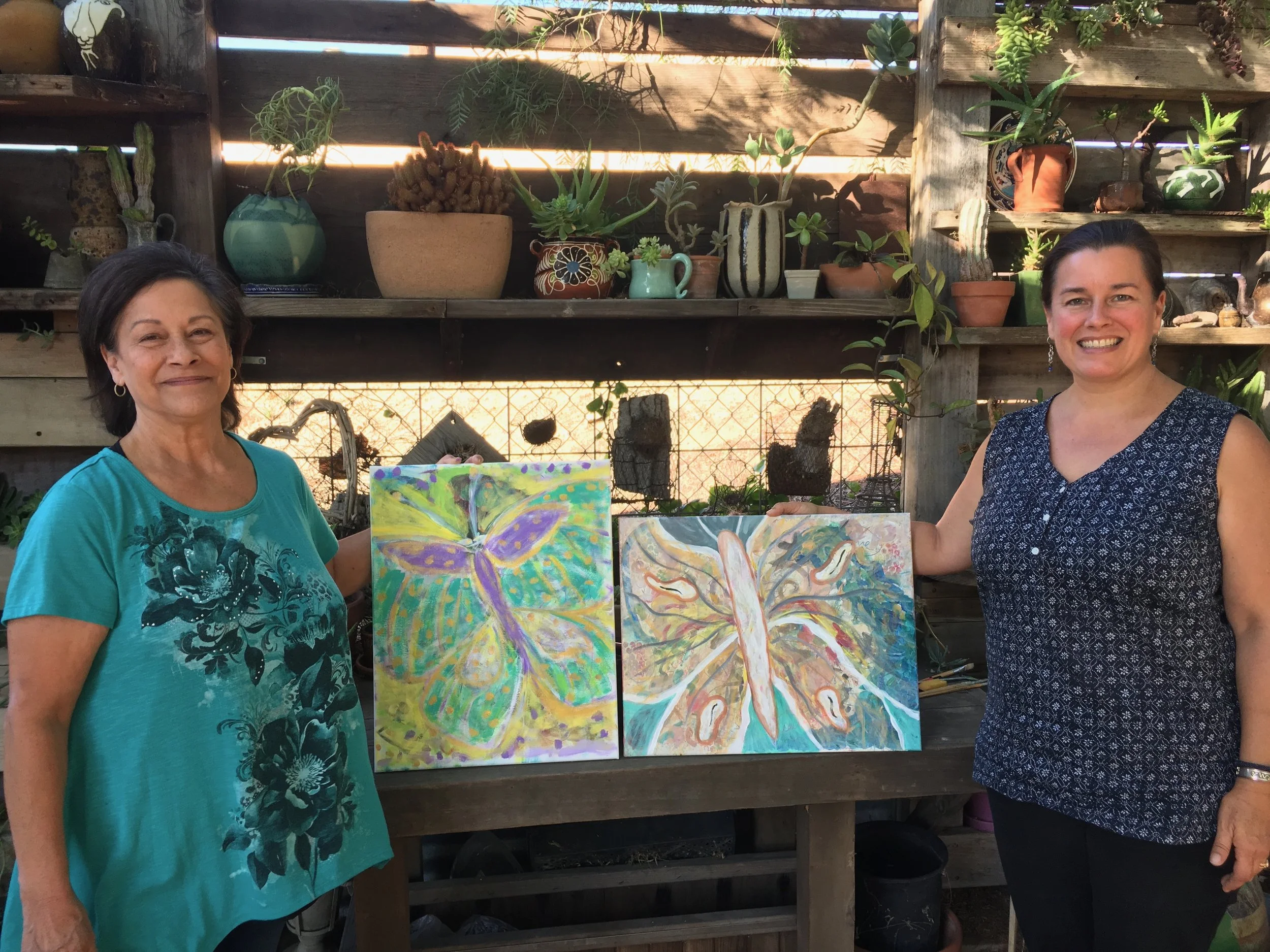 Two women standing in front of a wooden garden wall with shelves of potted plants and succulents, holding colorful paintings of butterflies and hands.