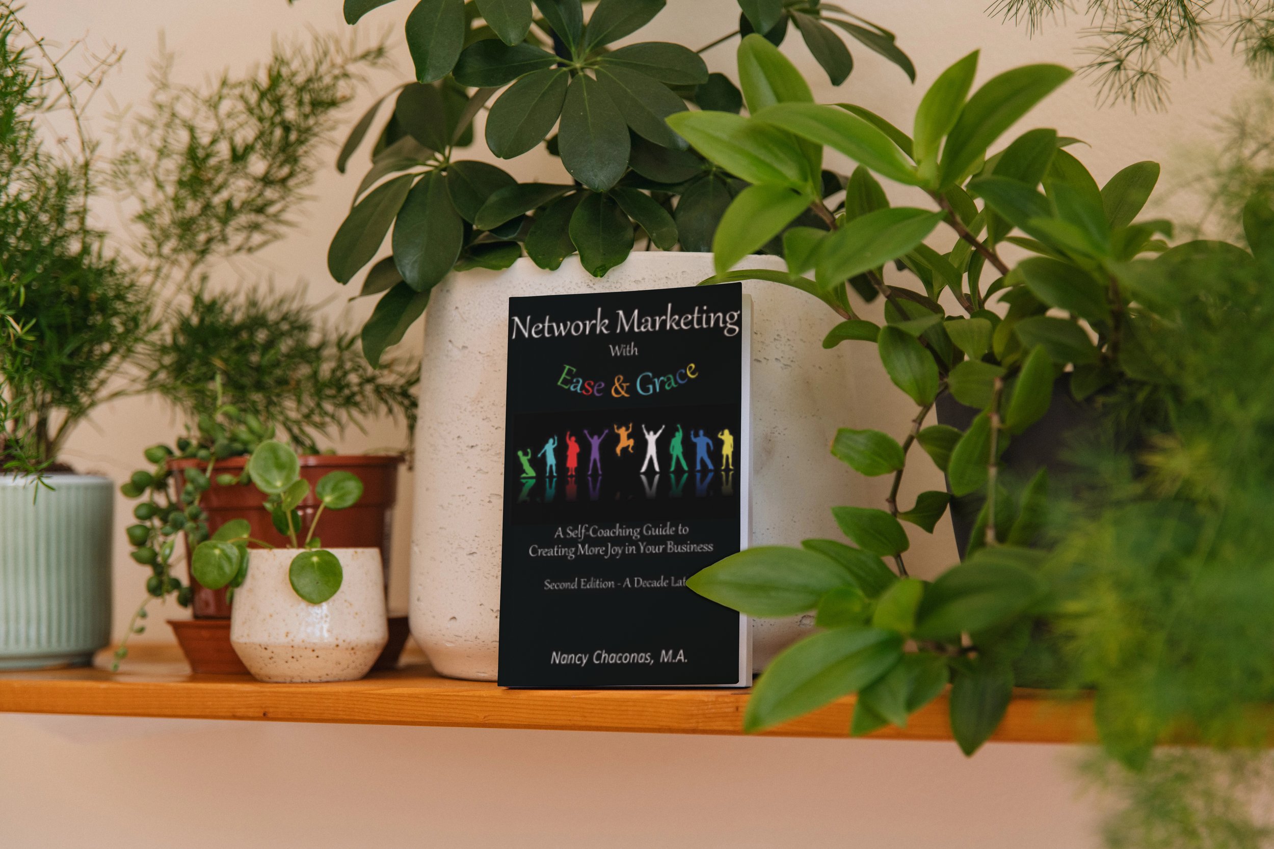Various potted plants on a wooden shelf, with a book titled 'Network Marketing with Ease & Grace' among them.