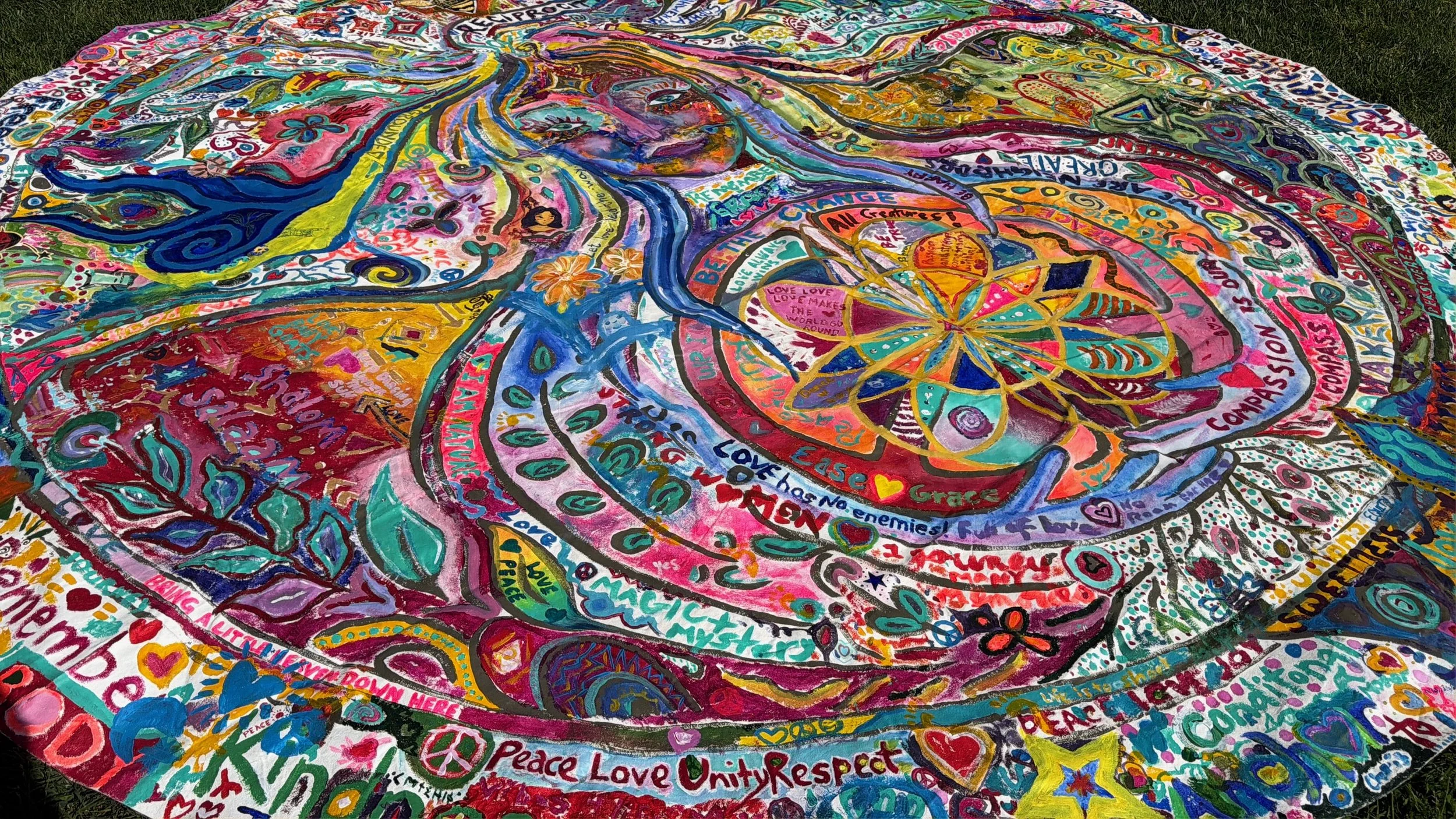 Colorful, large fabric with various messages, symbols, and drawings promoting love, peace, unity, kindness, and positivity, spread out on a grassy area.