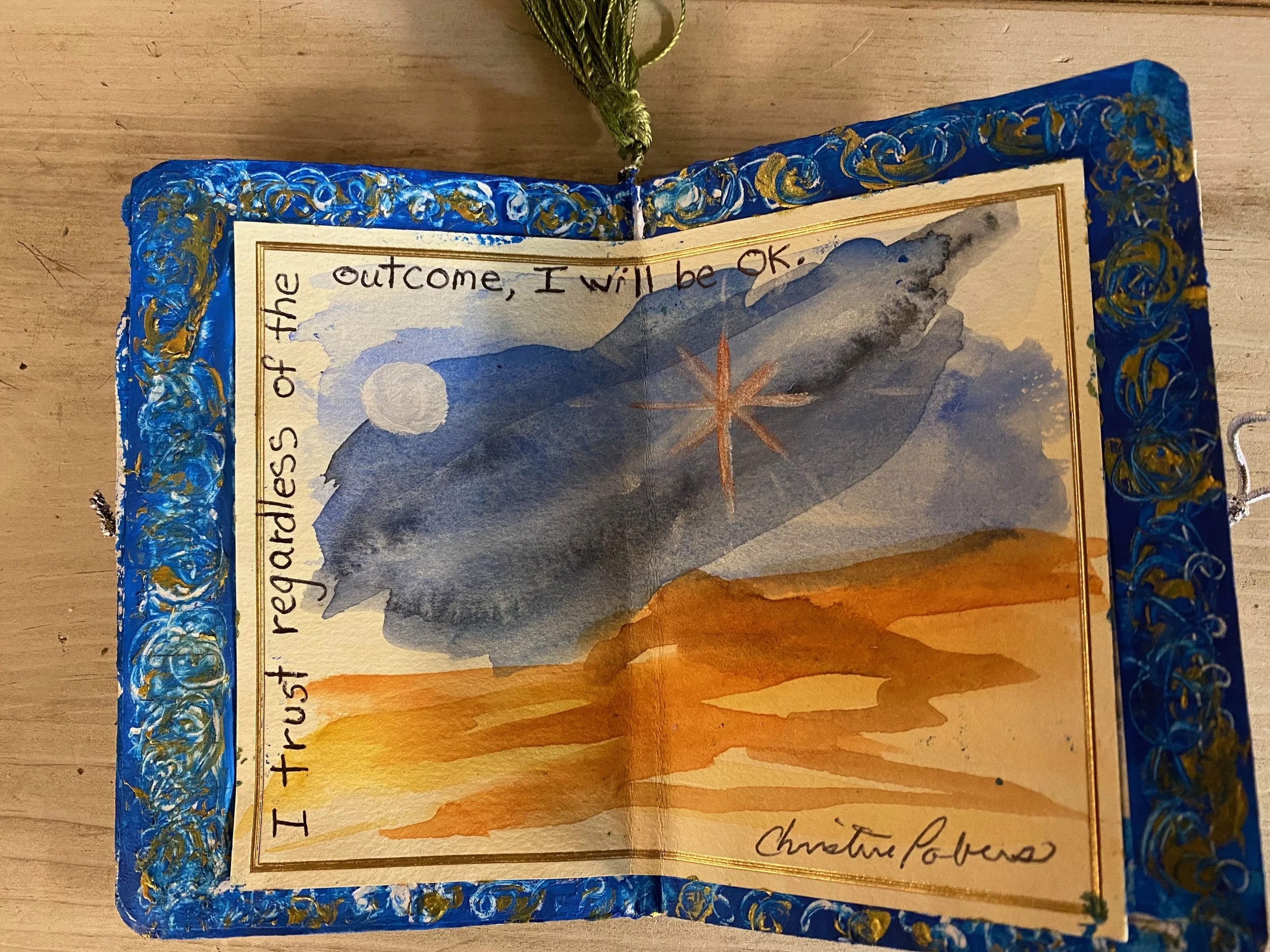 A decorated handmade card or picture frame with a blue, gold, and white border, featuring a watercolor scene of mountains, a sunset, and an orange star or sun. The scene includes handwritten text along the top and bottom edges, with part of it visibl