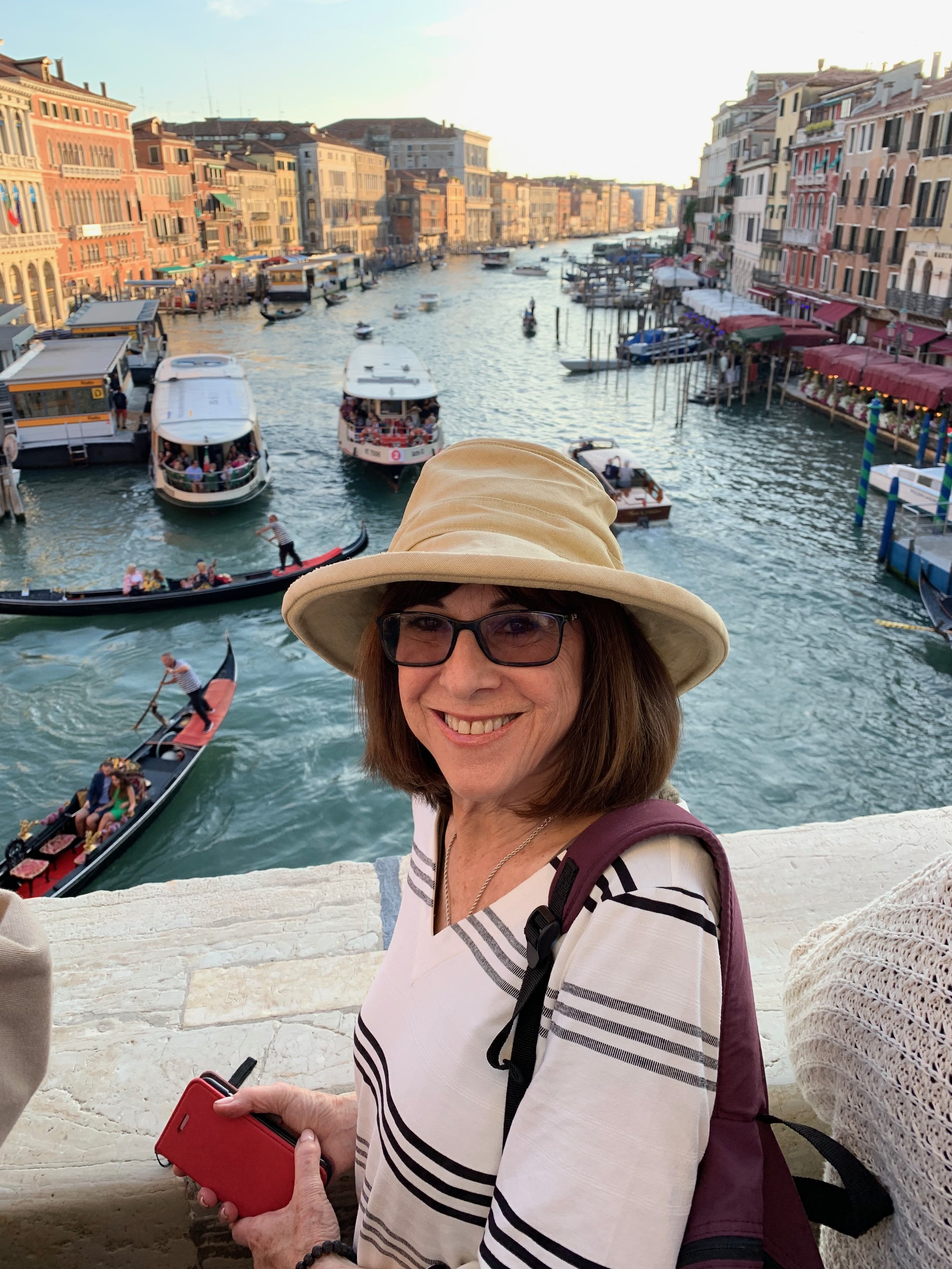 A woman with glasses smiling, wearing a beige hat with a wide brim and a striped shirt, taking a selfie with Venice's Grand Canal in the background. The canal is filled with boats, and there are colorful historic buildings lining the water.