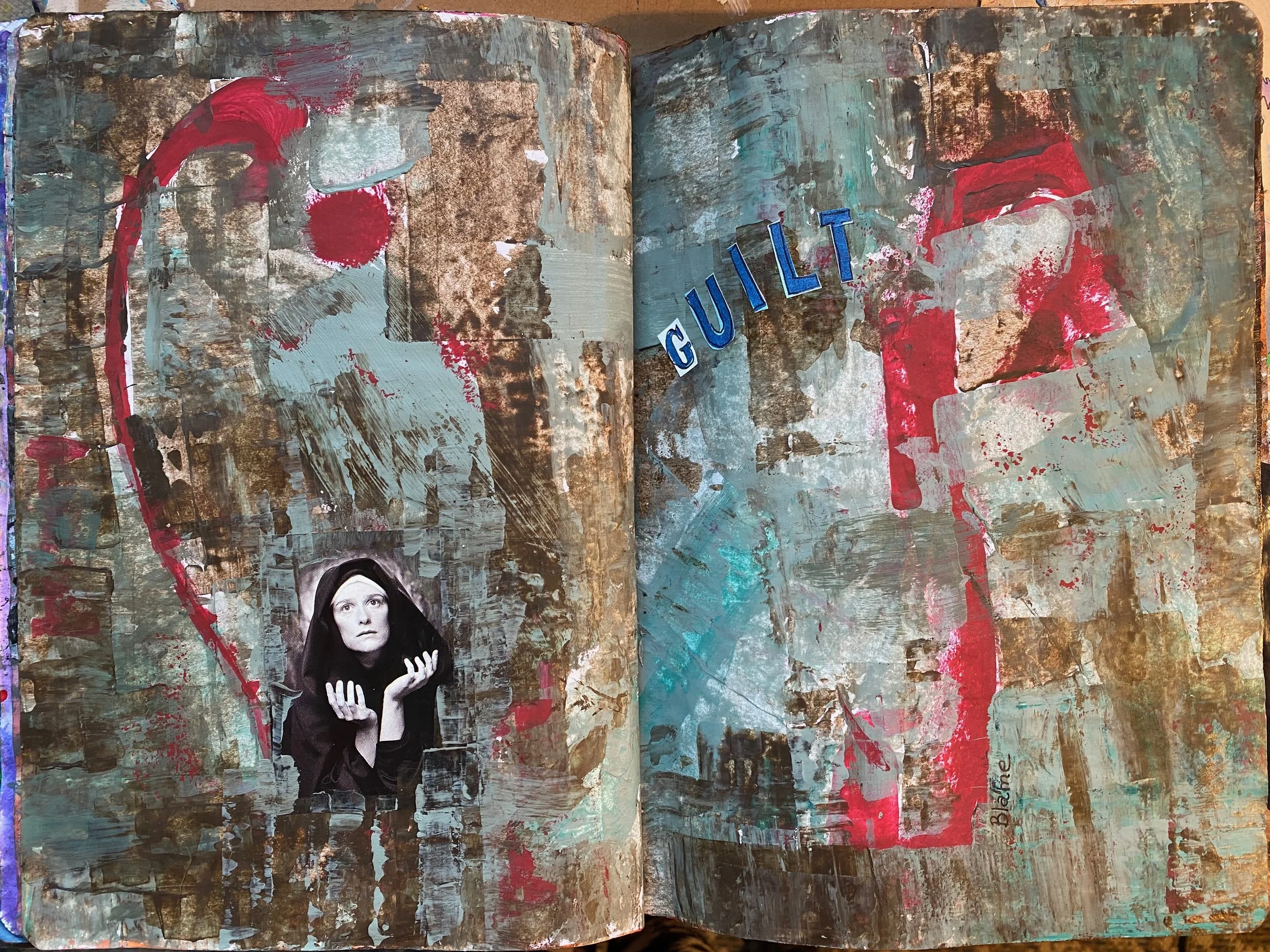 An open sketchbook with abstract, textured paint strokes in various colors including turquoise, gold, white, red, and black. The right page features the word "GUILT" in blue letters. The bottom left corner of the left page has a black-and-white cutou
