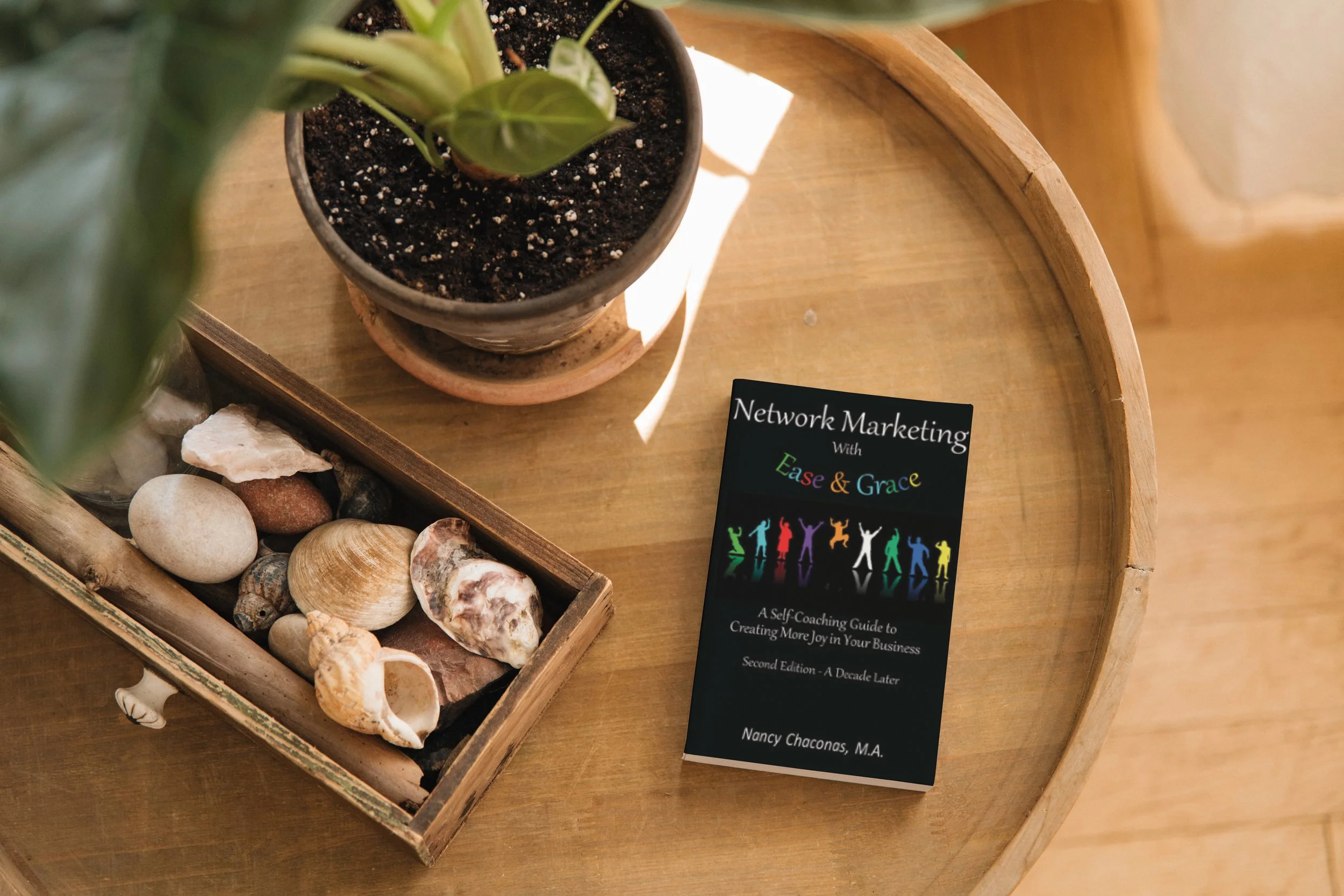 A wooden tray holding seashells, a small potted plant, and a business card for a self-coaching guide titled 'Network Marketing with Ease & Grace' by Nancy Chaconas, M.A.