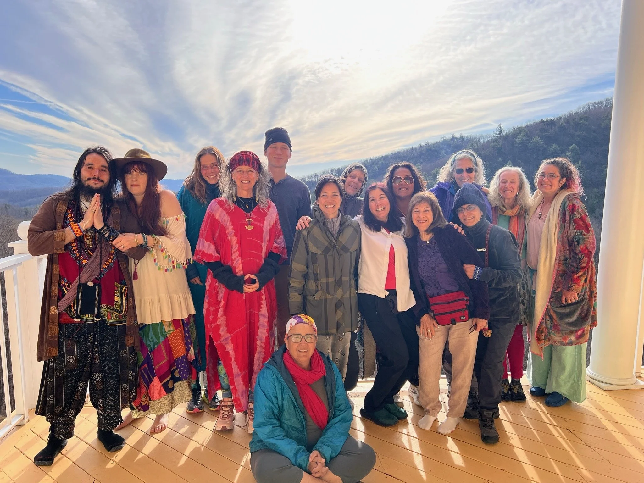 A group of people smiling together on a wooden deck outdoors under a partly cloudy sky, with mountains in the distance.
