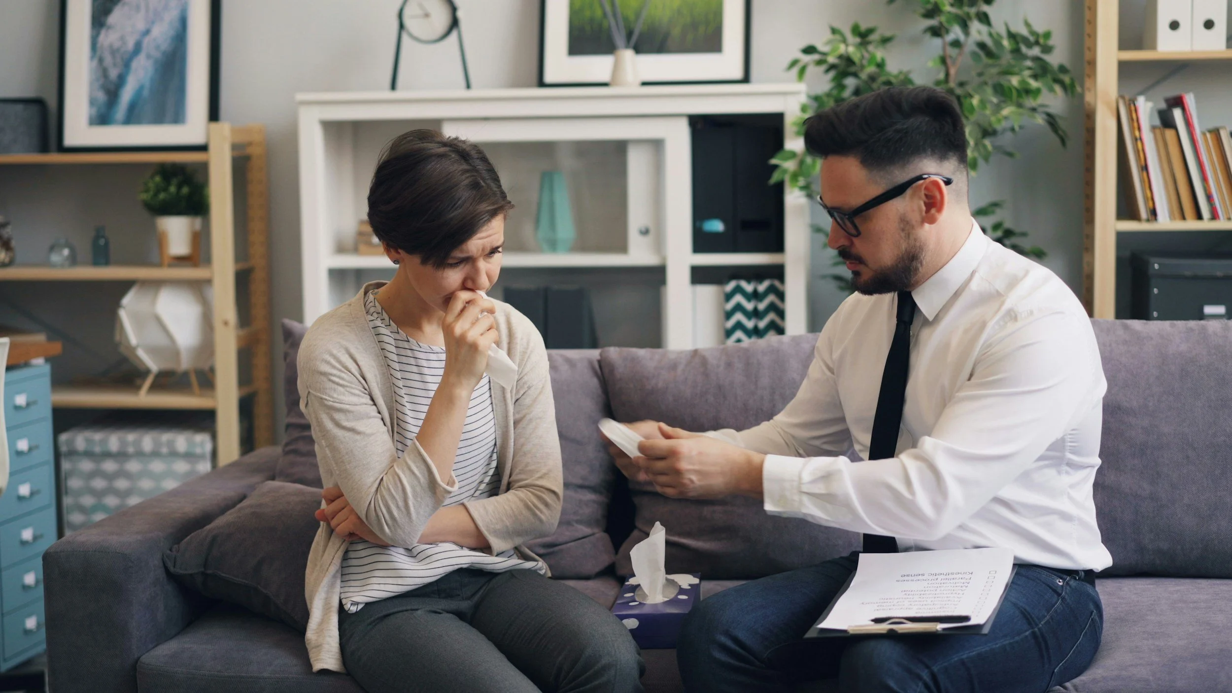 A woman with short dark hair wearing a striped shirt and beige cardigan sits on a couch with tears in her eyes, clutching her hand to her face, while a man with glasses and dark hair, dressed in a white shirt and tie, shows her a tissue and some papers, in a living room with picture frames and shelves in the background.