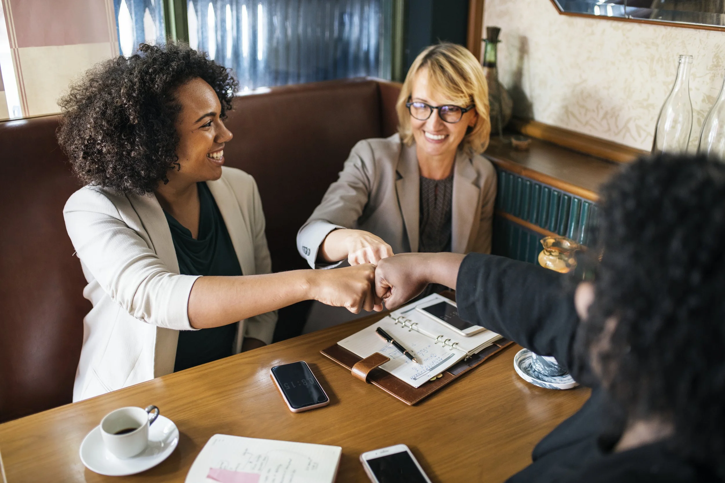 Two women smiling and shaking hands across a wooden table in a cozy cafe or restaurant, surrounded by notebooks, phones, and a cup of coffee.