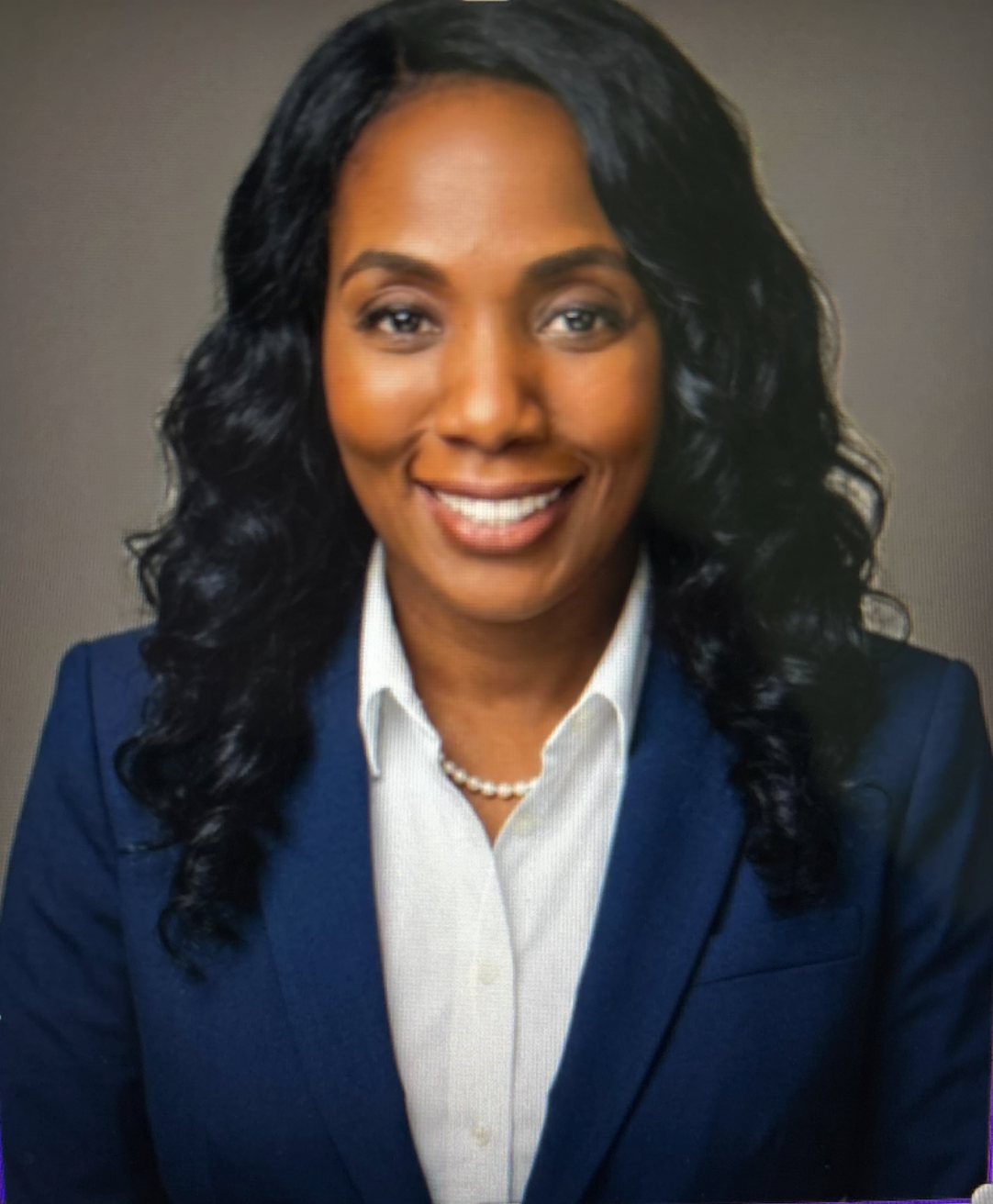 Headshot of a smiling woman with dark curly hair, wearing a navy blazer, white collared shirt, and pearl necklace, against a plain gray background.