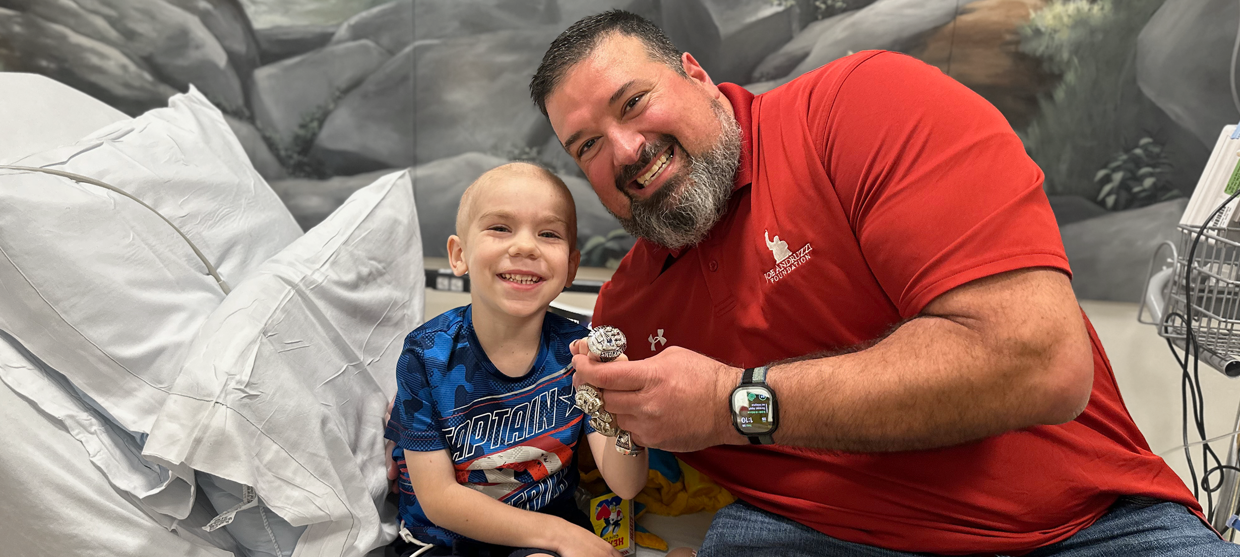 A young boy in a hospital bed holding a medal, smiling, with a large man in a red shirt sitting beside him, also smiling. The background shows hospital bed linens and a mural of rocks and plants.