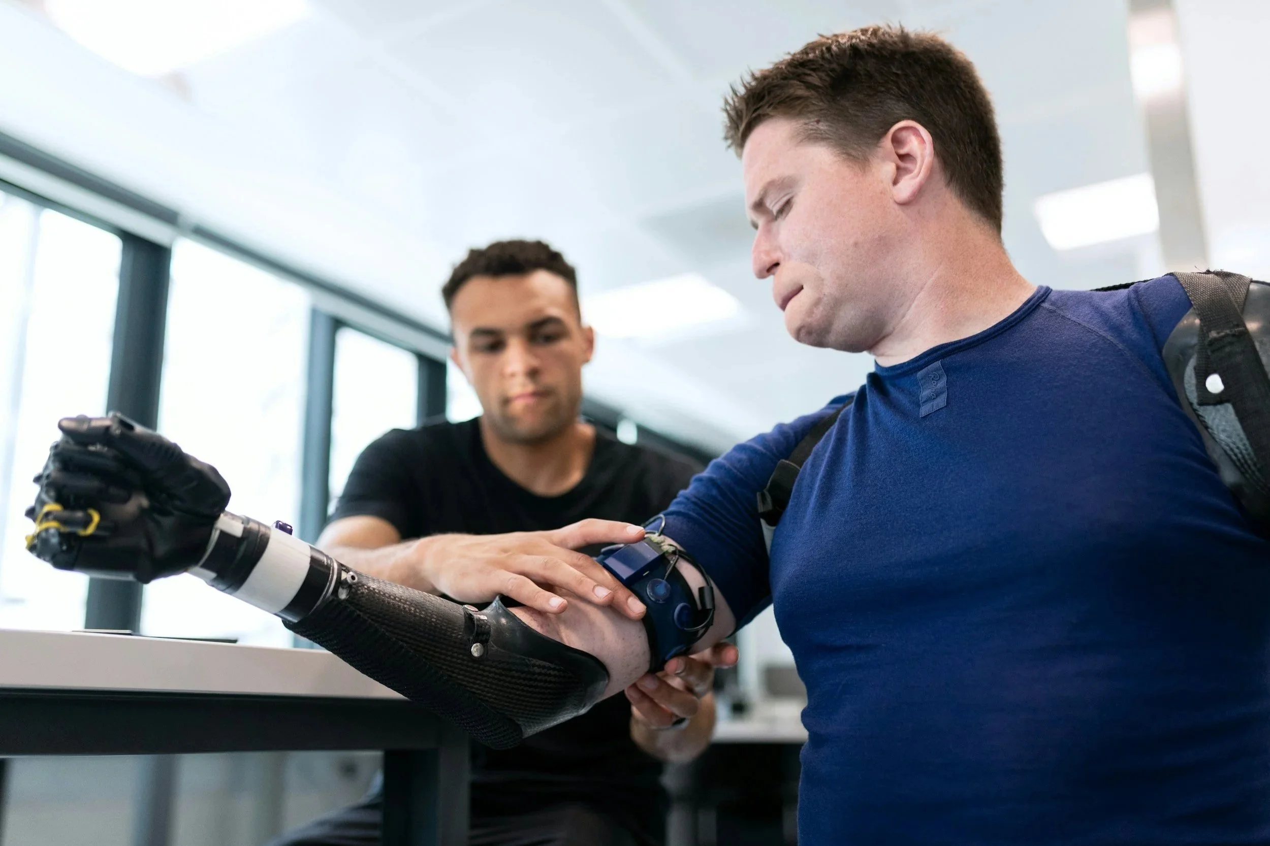 Two men, one with short brown hair and another with short dark hair, are in a modern indoor setting. One man is assisting the other with a robotic arm, likely in a medical or technological context. The man in blue is focused on the robotic arm, which is being handled by the man in the black shirt. Bright natural light streams in through large windows in the background.