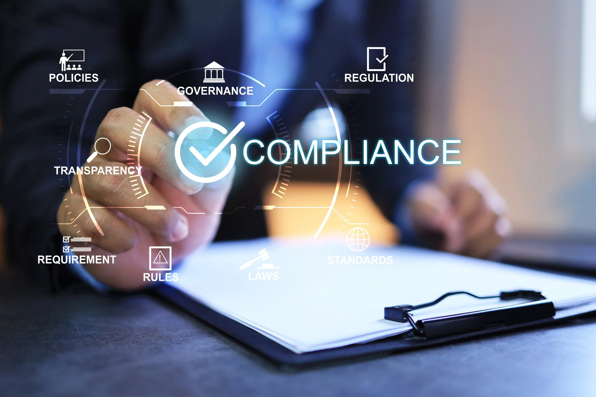 A person is pointing at a futuristic digital interface with words and icons related to compliance, policies, governance, regulations, requirements, laws, rules, and standards, over a desk with a clipboard and pen.